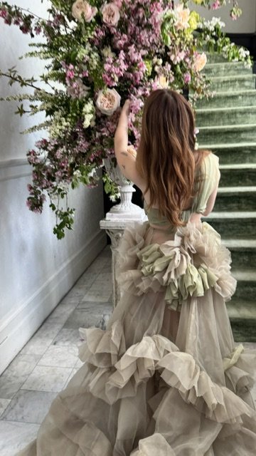 A woman with long, wavy brown hair wearing a light beige dress with ruffles standing beside a large floral arrangement on a staircase.