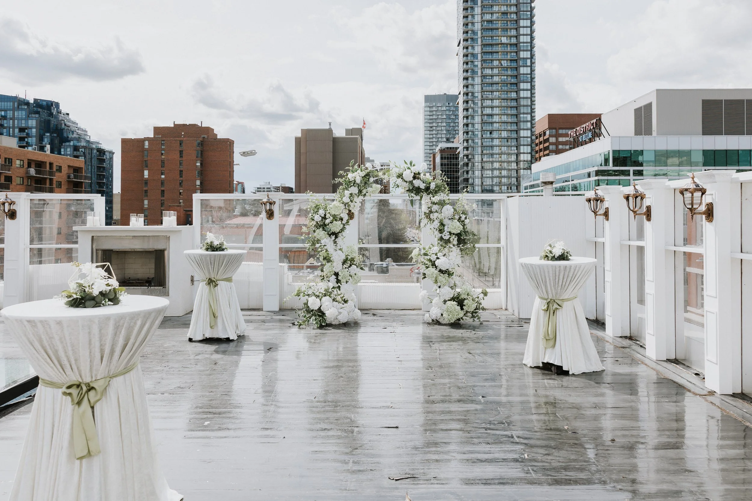 Decorated outdoor rooftop wedding ceremony space with floral arrangements and tables, surrounded by city skyscrapers under a cloudy sky.