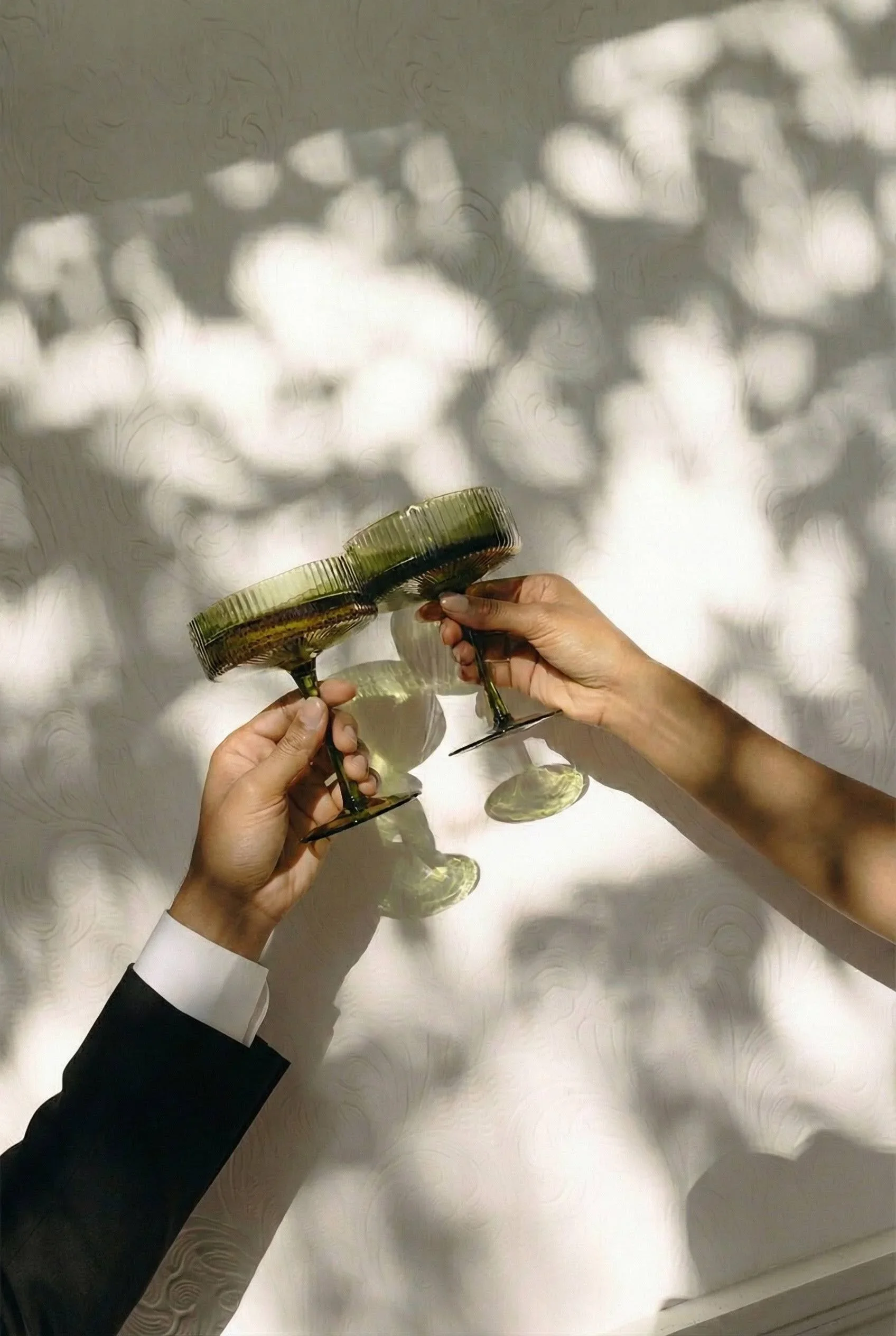 Two hands holding green glasses with reflections casting shadows on a white textured surface.