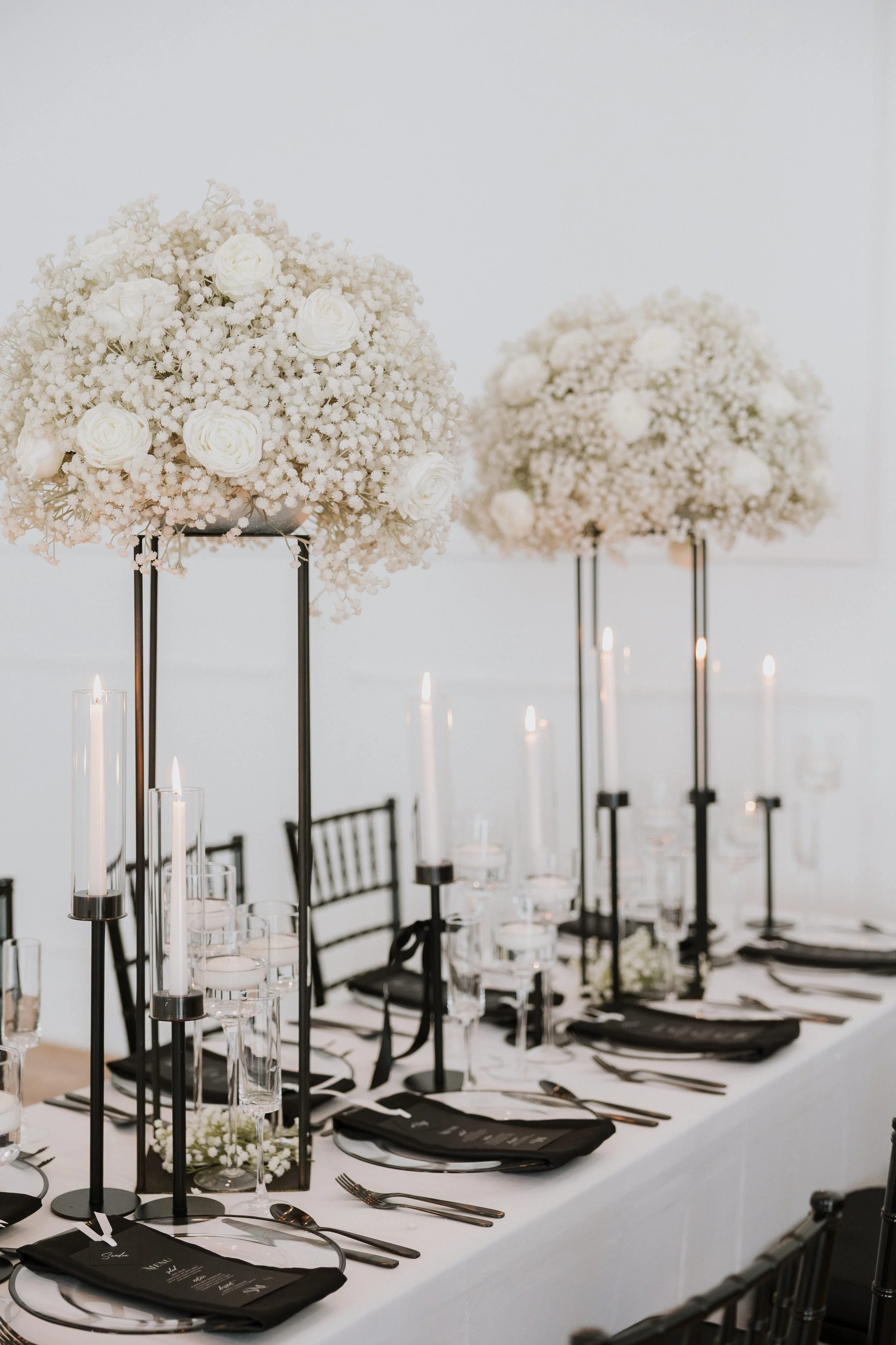 Elegant wedding table setup with tall floral centerpieces of white roses and baby's breath, black chairs, black napkins, candles in glass holders, and place settings with black menus.