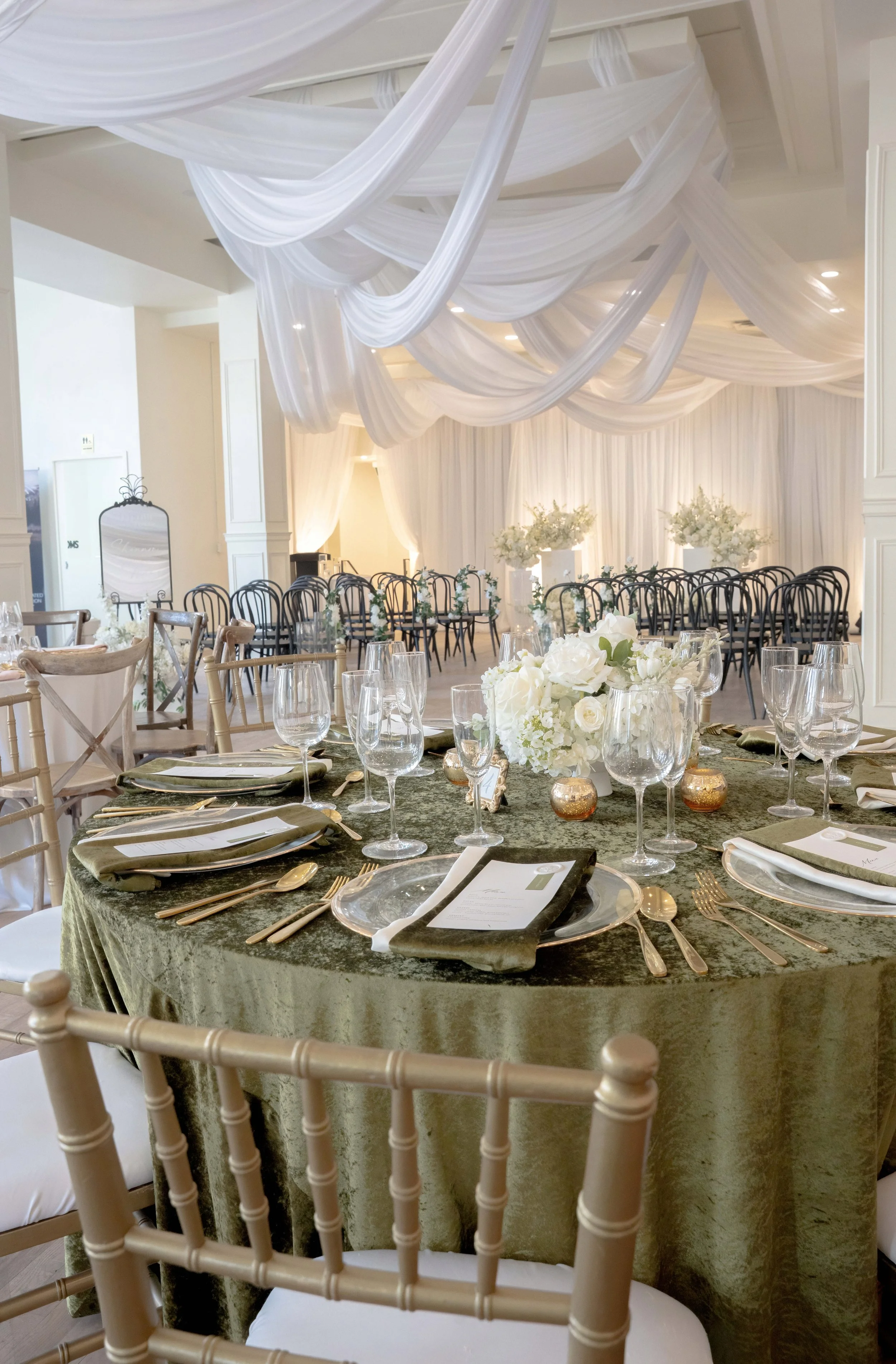 A round table covered with a green velvet tablecloth, set with plates, silverware, and glasses for a formal event. A floral centerpiece features white flowers, and gold votive candle holders are scattered around. In the background, there are rows of 