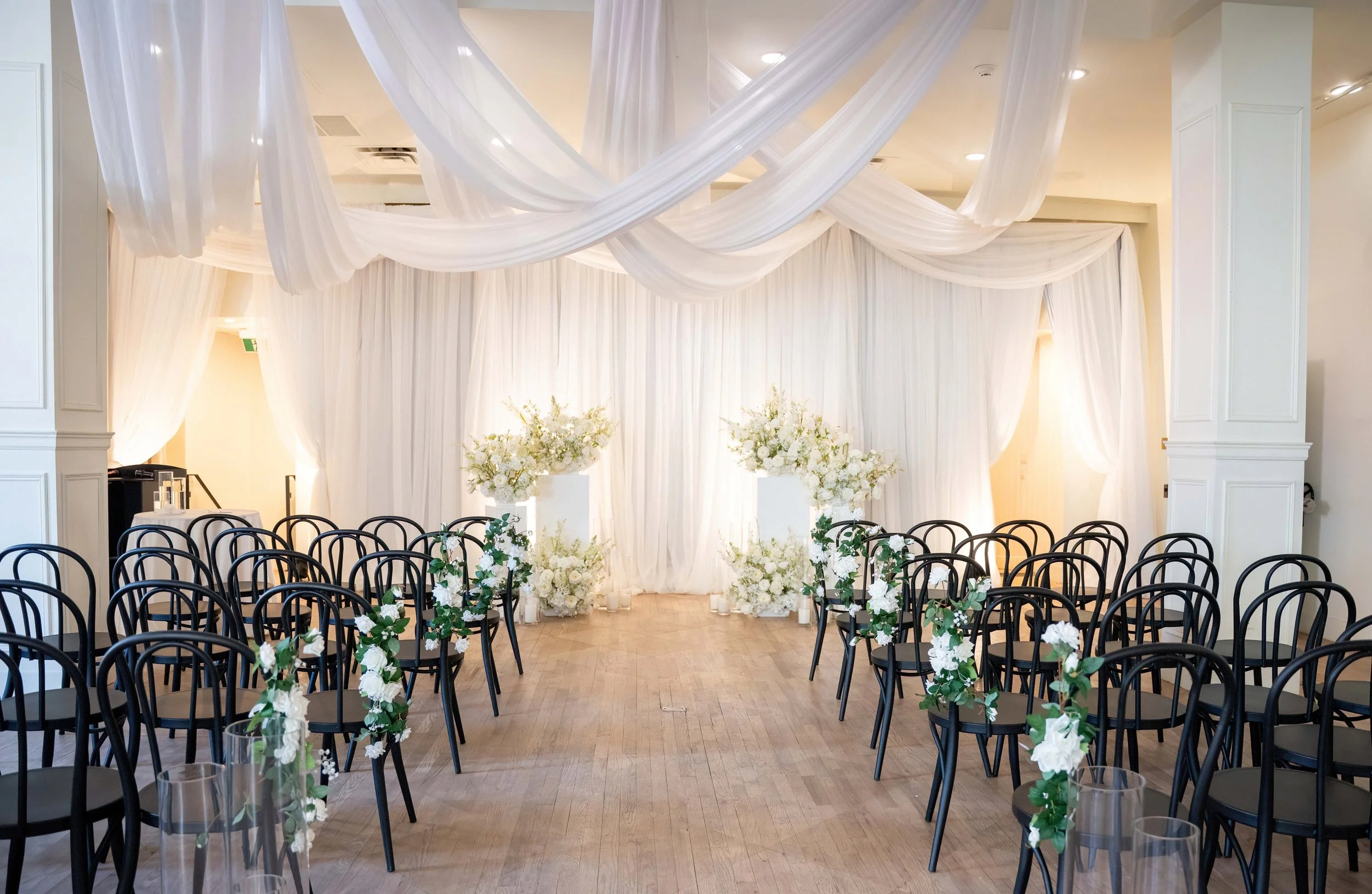 A wedding ceremony setup with rows of black chairs decorated with white flowers on the aisle, white drapes and floral arrangements at the altar, and sheer white fabric swags hanging from the ceiling in a bright, elegant room.