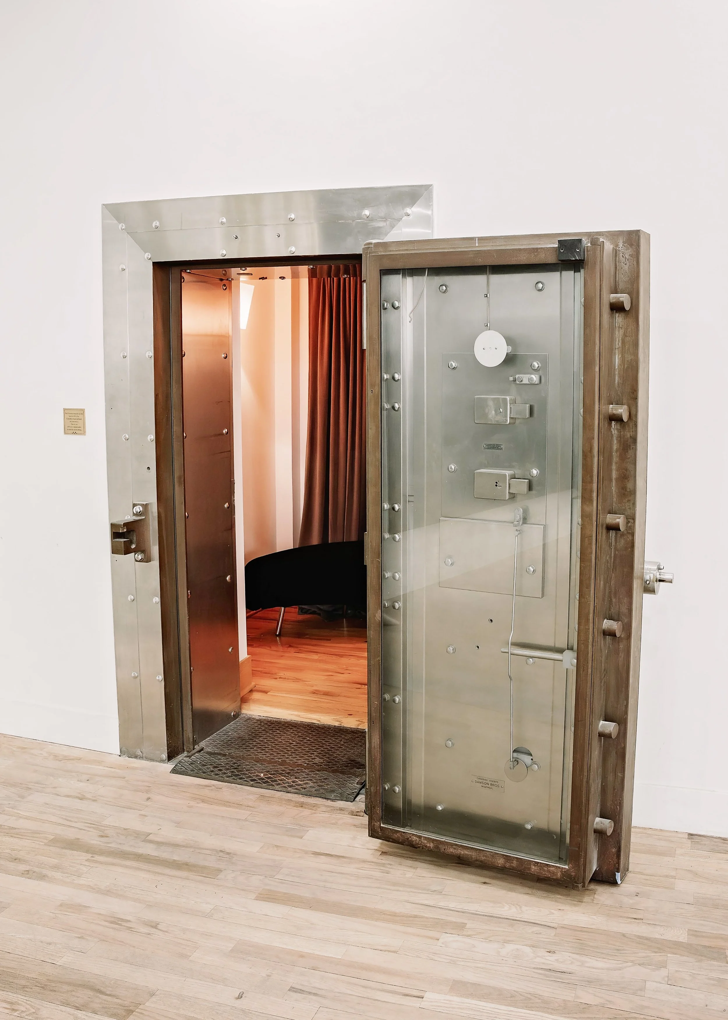 A large, open vault door with a combination lock and locking mechanisms inside a room with white walls and wooden flooring.