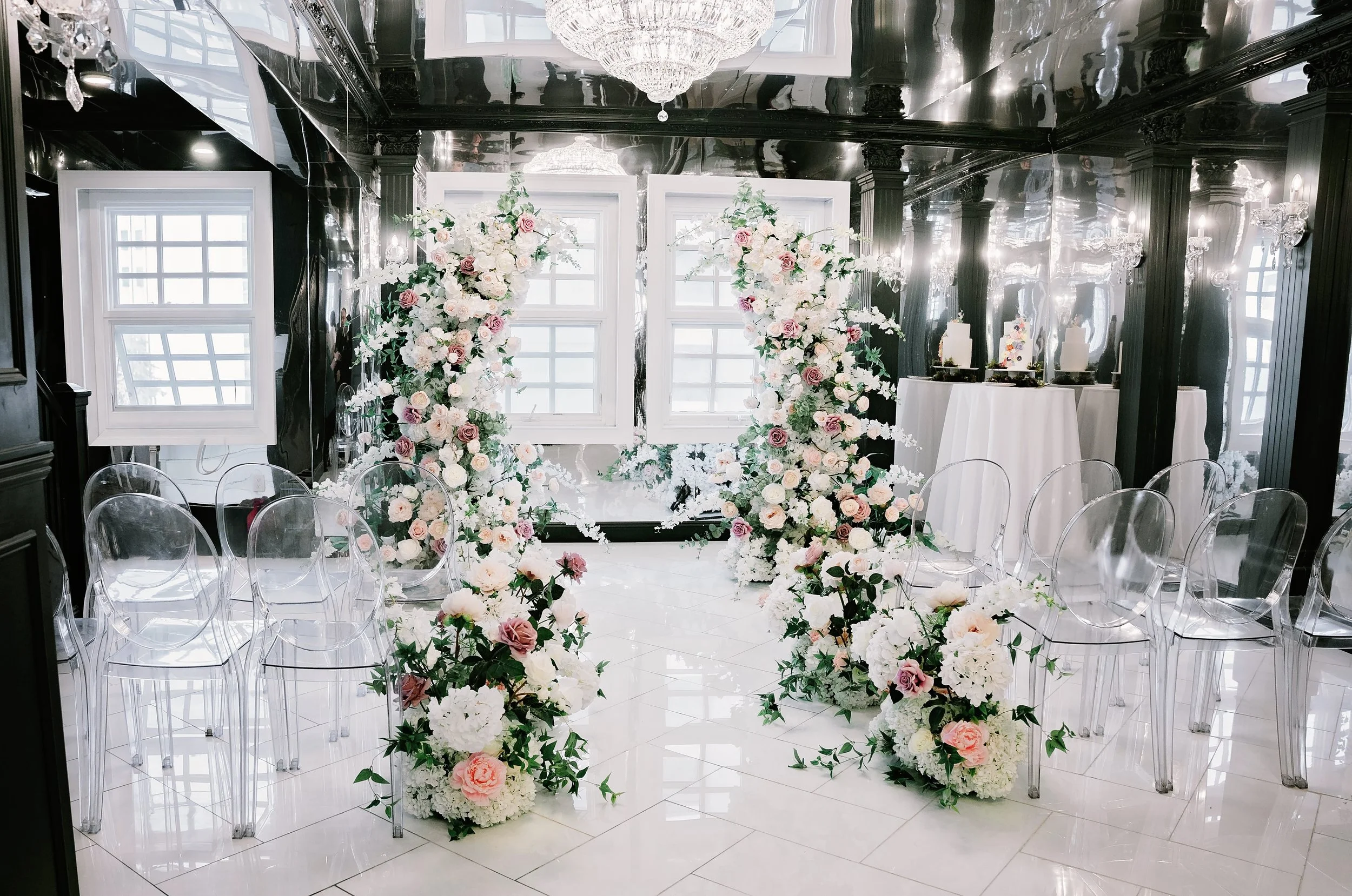 Elegant wedding or event setup with tall floral arrangements of white and pink roses and white flowers, clear acrylic chairs, and white-topped tables in a room with black glossy walls, white window frames, and a chandelier.