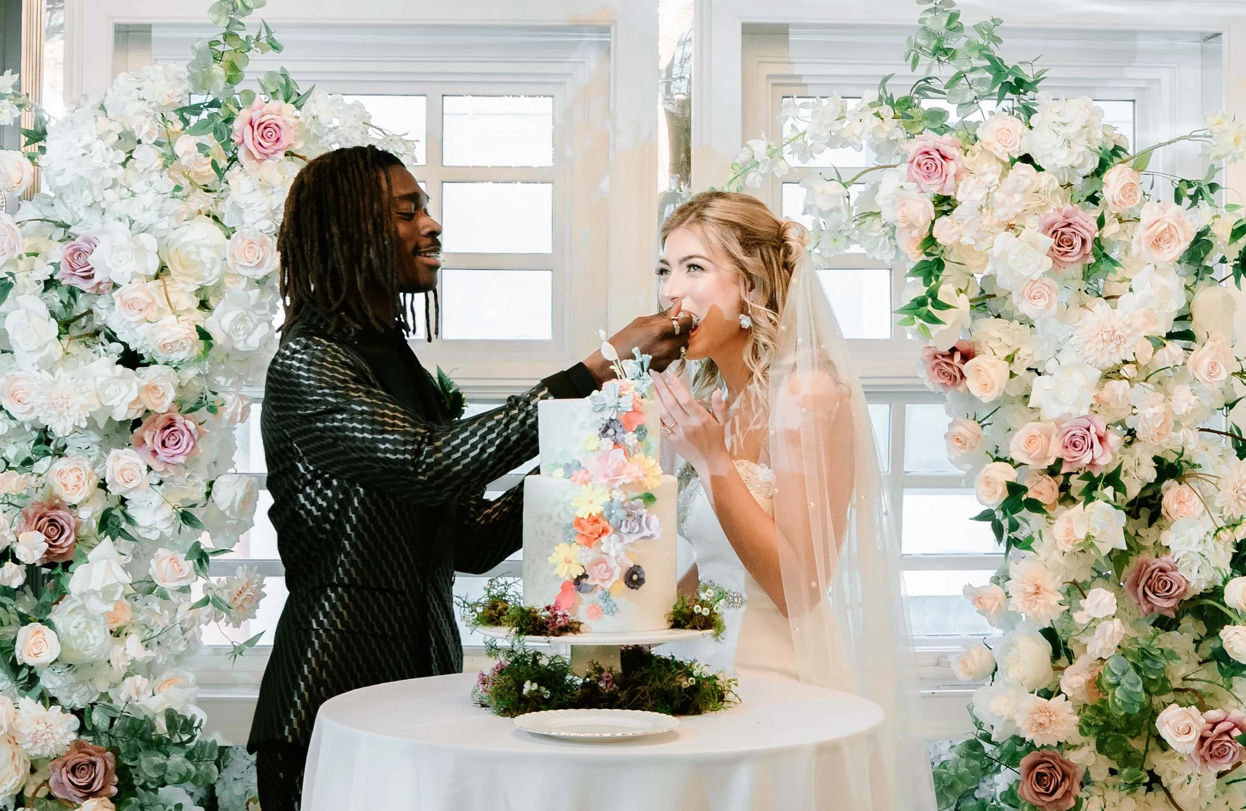 A wedding celebration where a man is feeding a woman wedding cake in front of floral arrangements with white and pink roses.