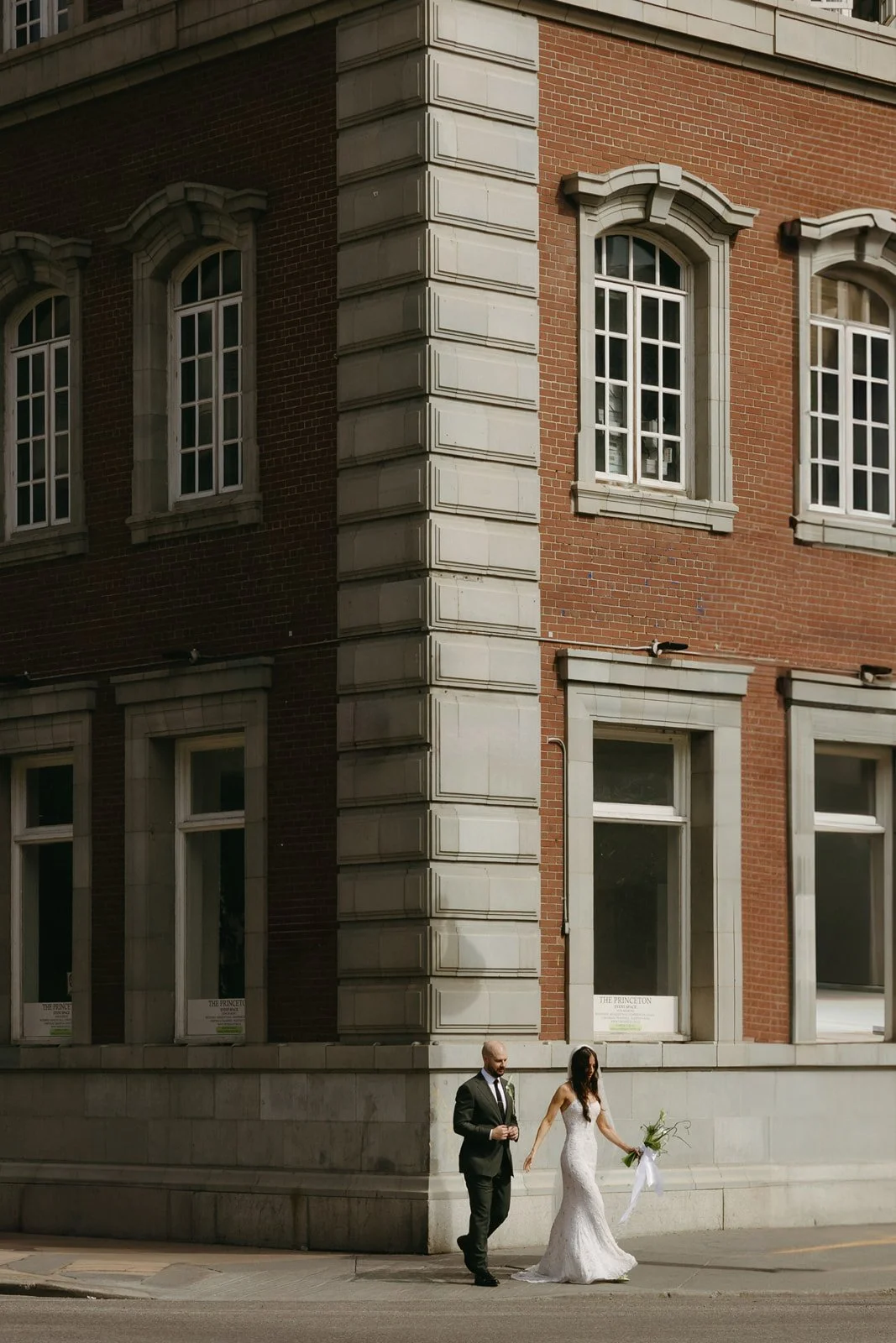 A bride and groom walk hand-in-hand on a city street in front of a brick building with large windows, the bride holding a bouquet of flowers.