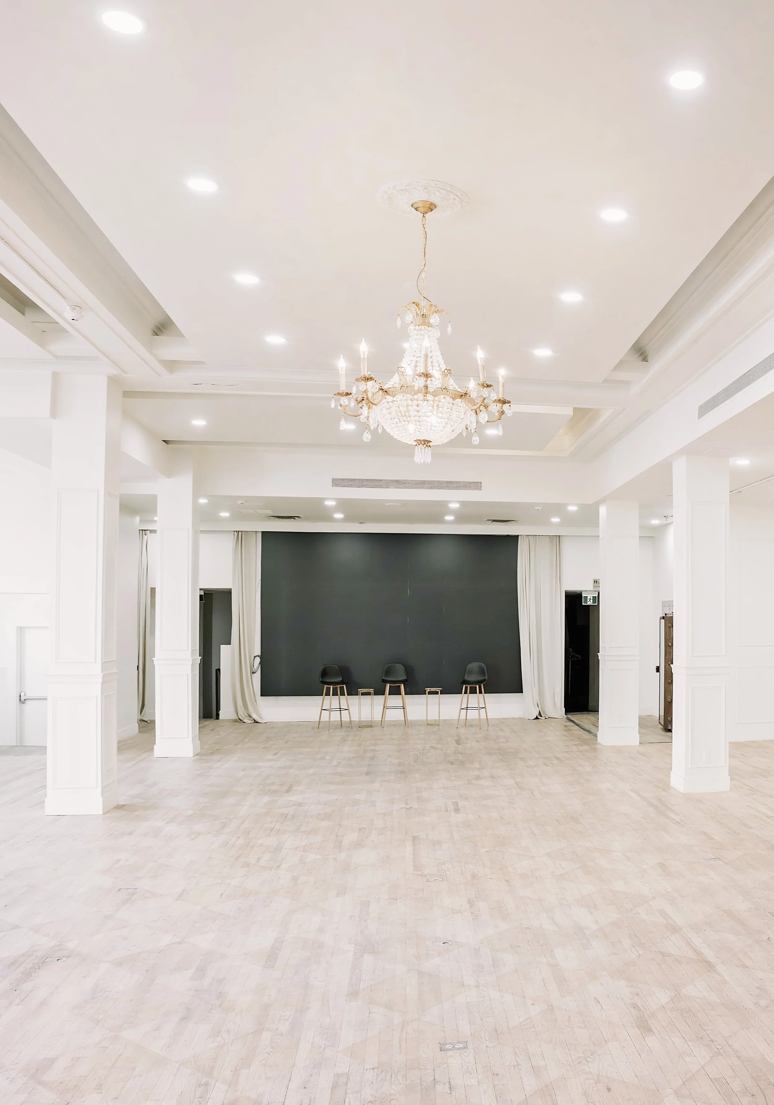 An empty, elegant event hall with white walls, a high ceiling, and a large chandelier. The floor is light-colored wood, and there are three black chairs and three small tables in front of a dark gray backdrop. The hall has drapes on the sides and cei