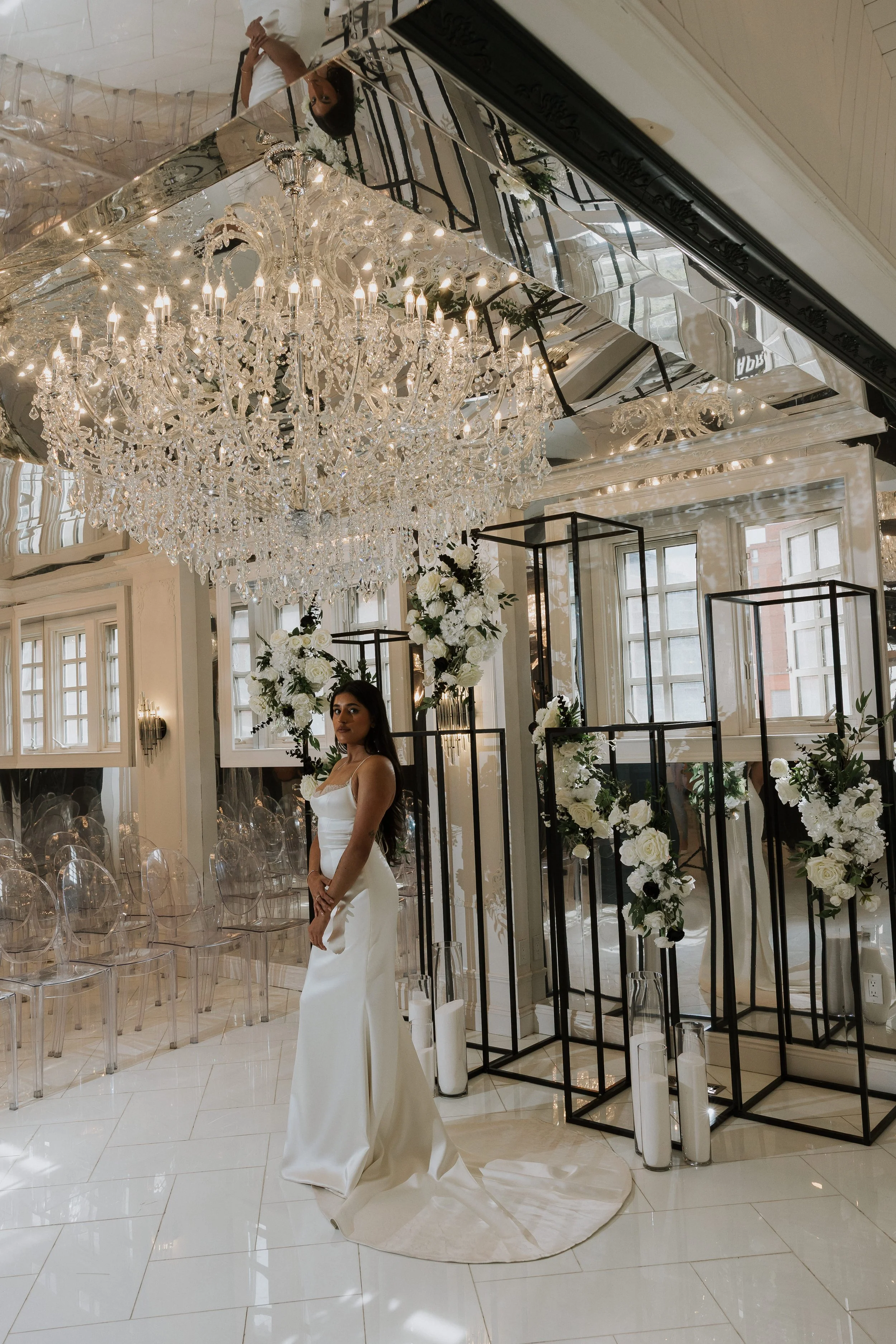 Interior of an elegant venue with a large crystal chandelier, floral arrangements, and a woman in a white gown standing on a white circular rug.