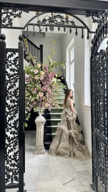 A woman in a beige gown standing in front of a staircase with black and white railing, framed by ornate black iron gates and a large floral arrangement on a pedestal.