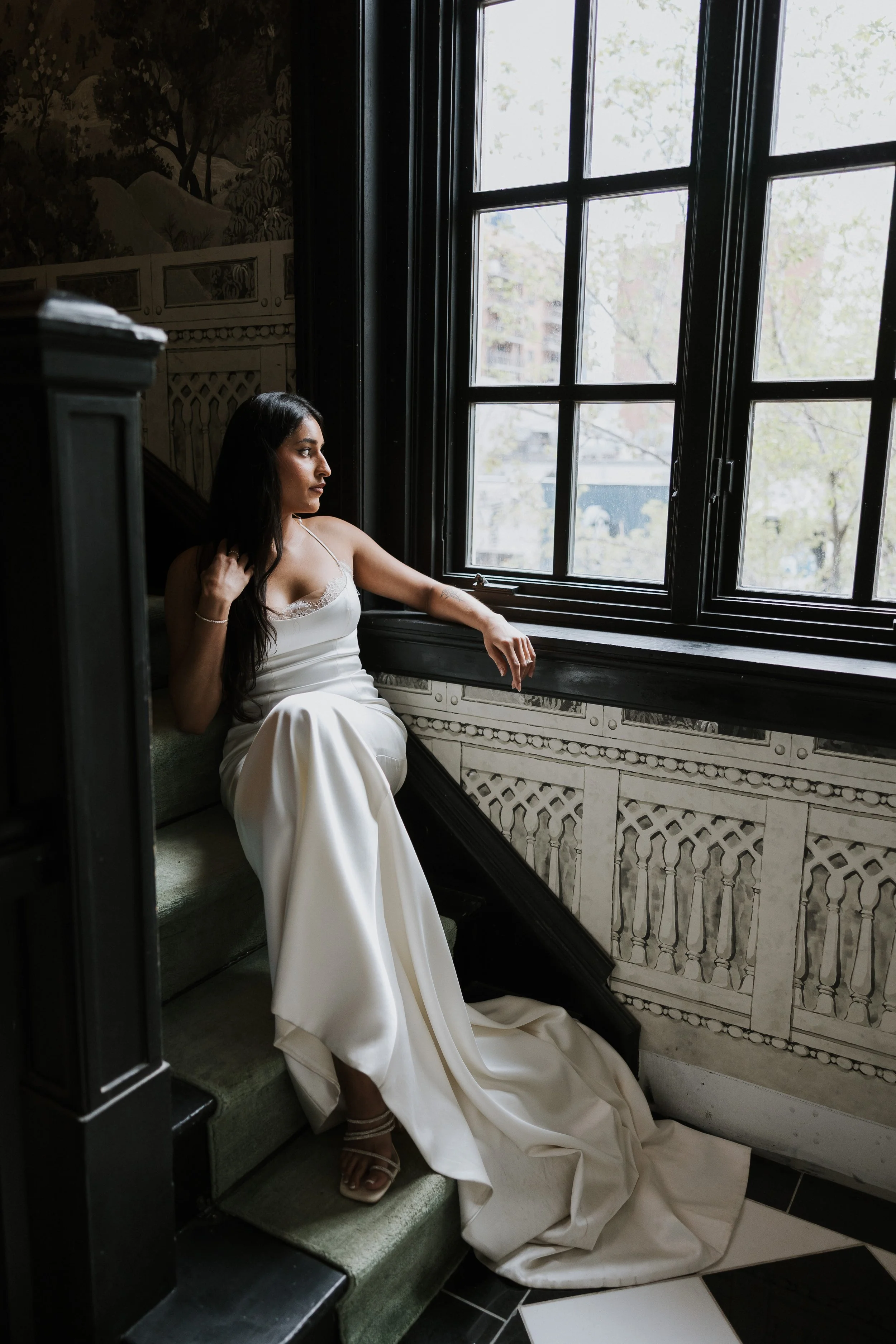 A woman with long dark hair in a white dress sitting on a staircase near a large window, looking outside.