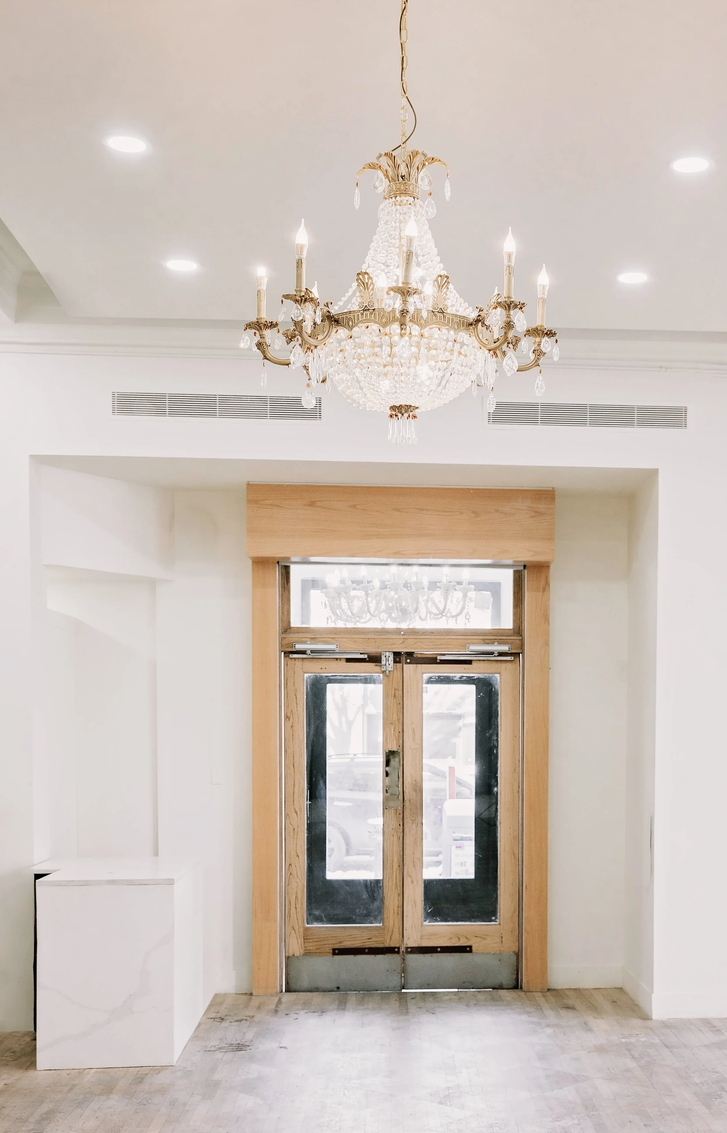 Interior view of a room with a crystal chandelier hanging from the ceiling, double wooden doors with glass panes, and white walls. There are recessed lights in the ceiling and a white marble pedestal on the left side.