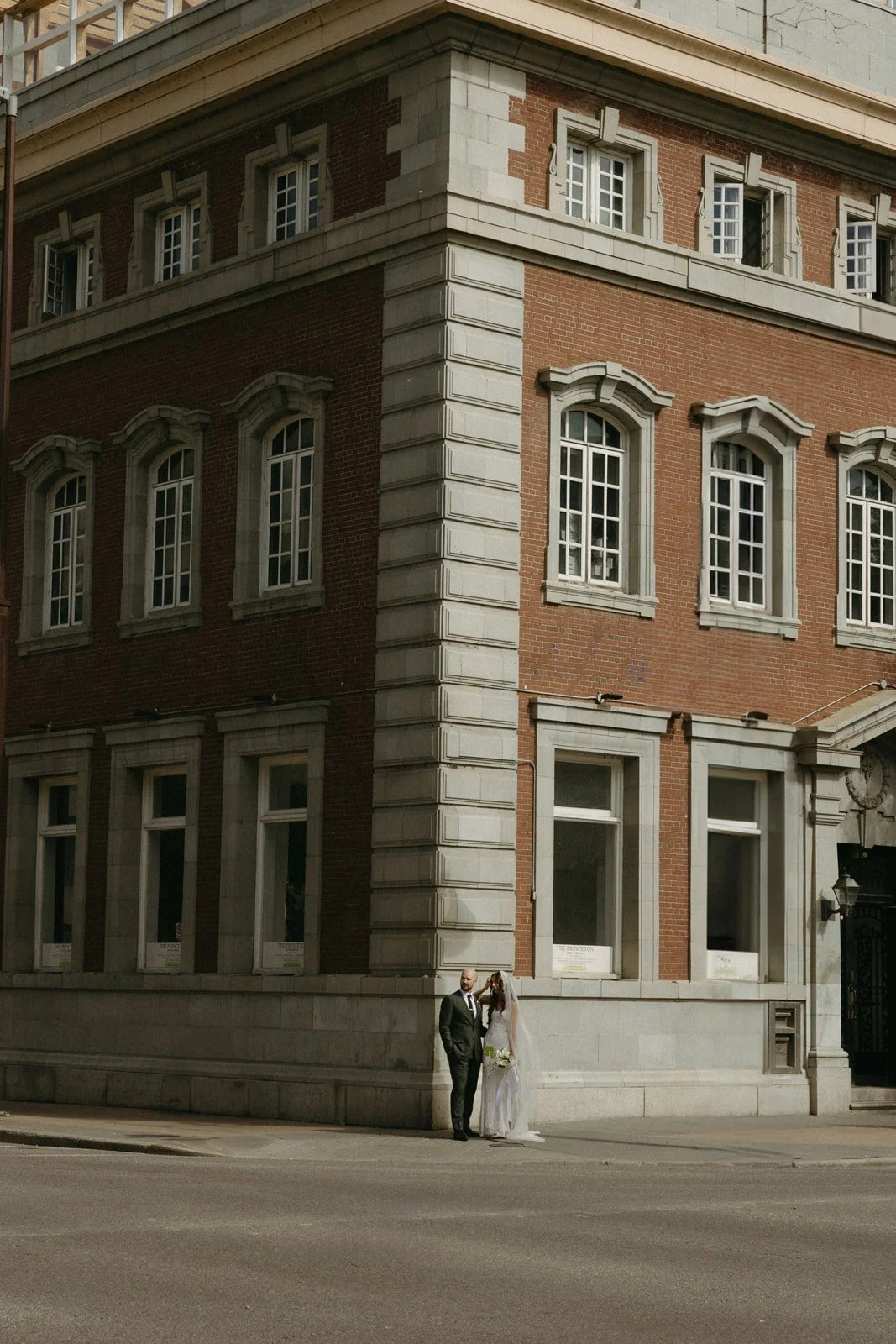 A man and woman dressed in wedding attire standing on the sidewalk in front of a large brick building with multiple large windows.