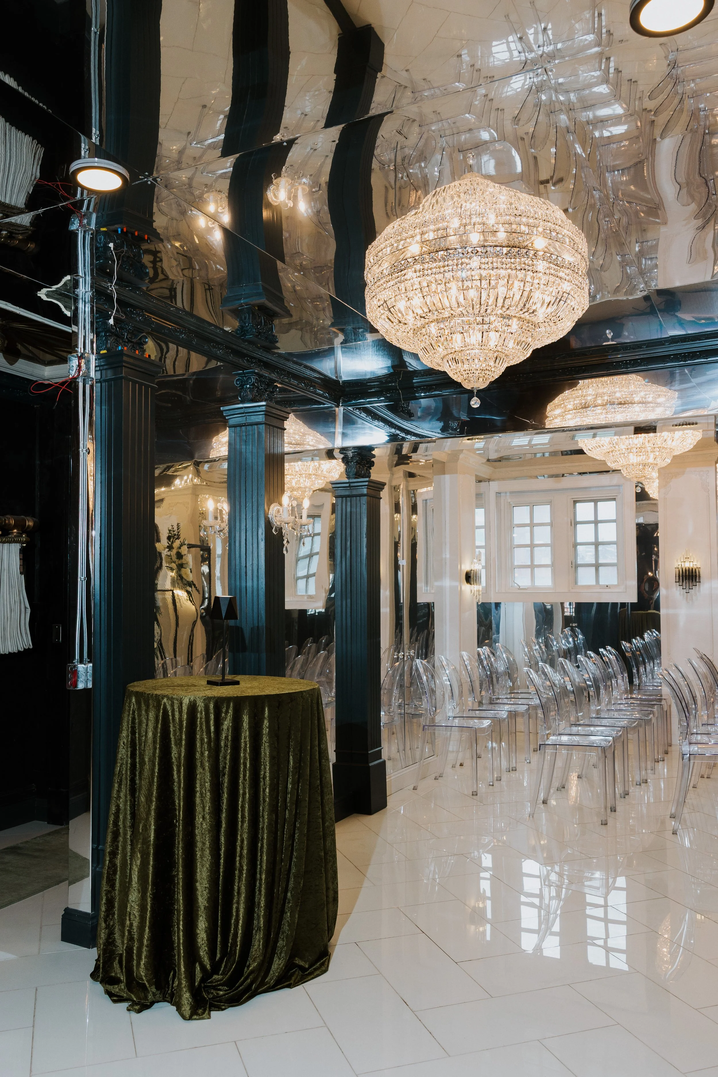 A luxury interior dining area with three large crystal chandeliers hanging from a mirrored ceiling. There are black decorative columns, clear acrylic chairs arranged in rows, and a round table with a green tablecloth in the foreground. Large windows with grid patterns are visible in the background.