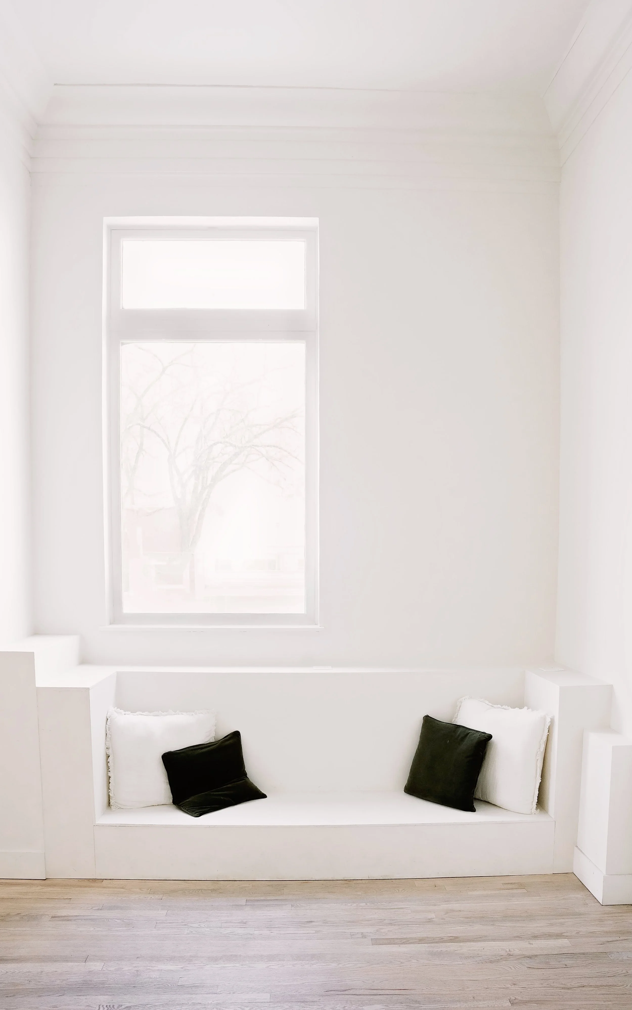 Contemporary white reading nook with a large window, built-in seating area, two white and two black throw pillows, and light-colored hardwood floors.