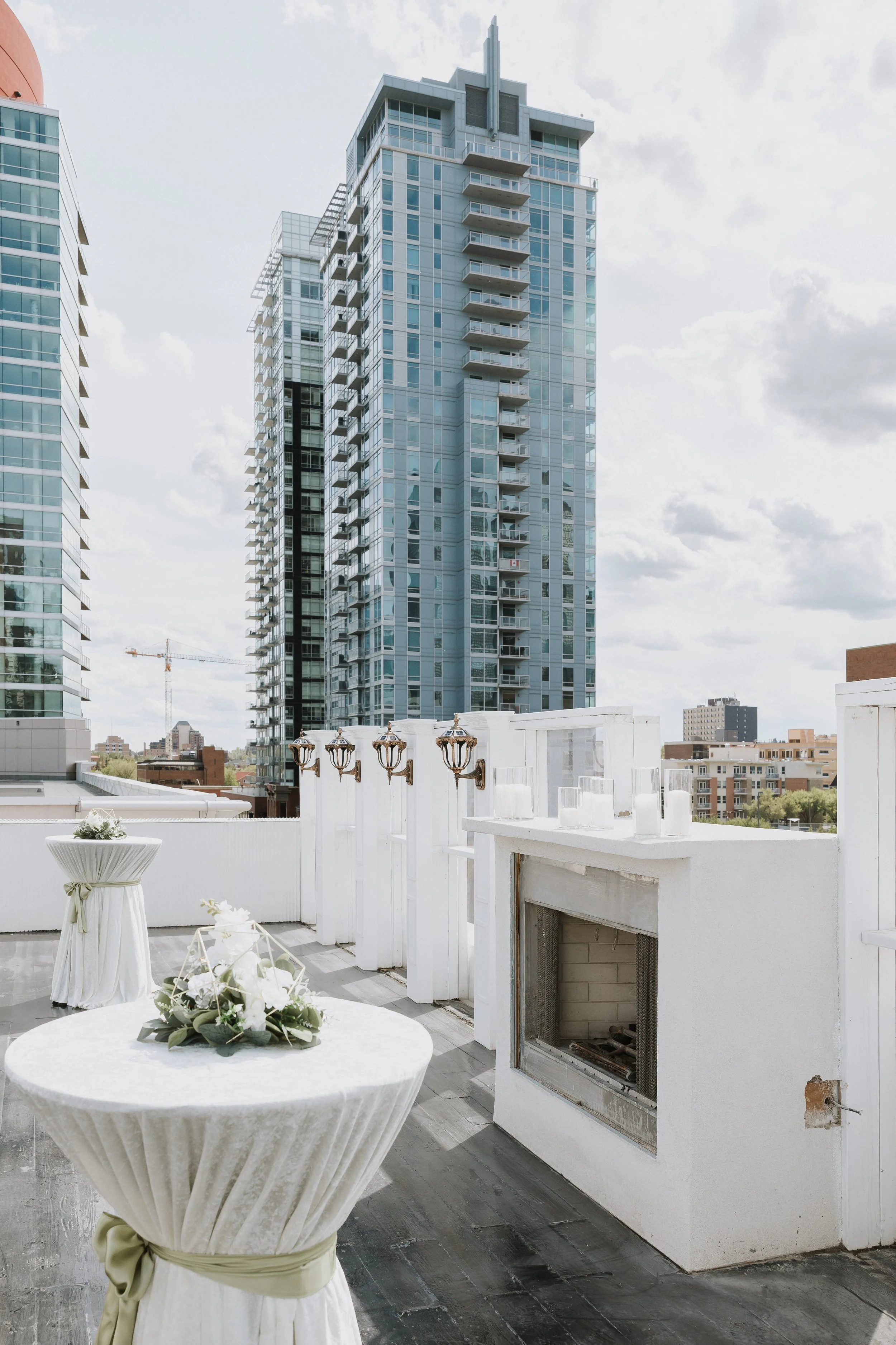 A rooftop outdoor space with a white table adorned with a floral centerpiece, surrounded by white draped chairs, with tall modern glass buildings in the background under a cloudy sky.