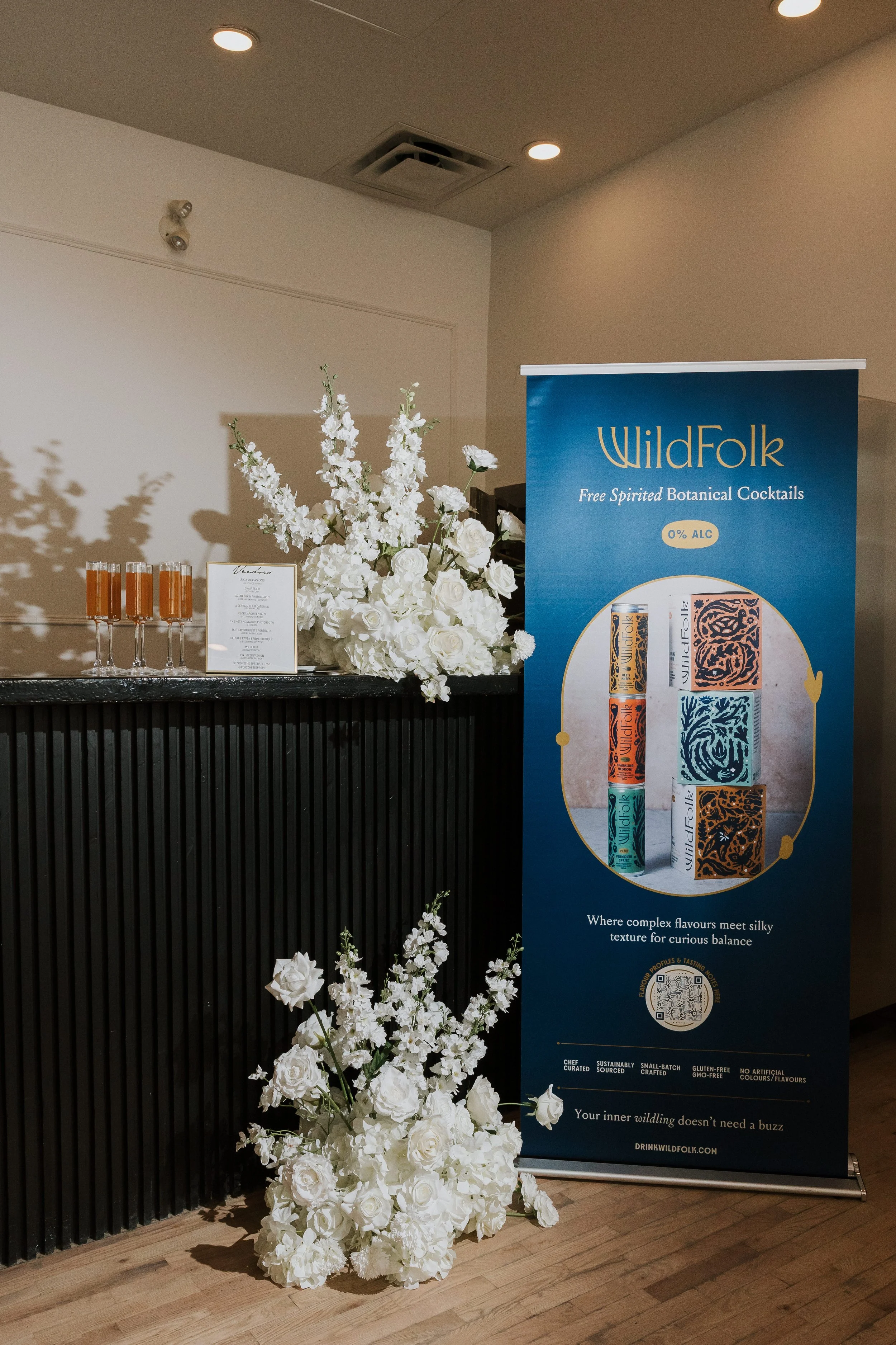 A display at an event for WildFolk beverages. The setup includes a tall arrangement of white flowers at the base, a large floral centerpiece with white roses and other white blooms, a menu or product description on a white framed board, four tall glasses filled with a reddish-orange drink, and a blue vertical banner advertising WildFolk botanical cocktails with a description and a QR code.