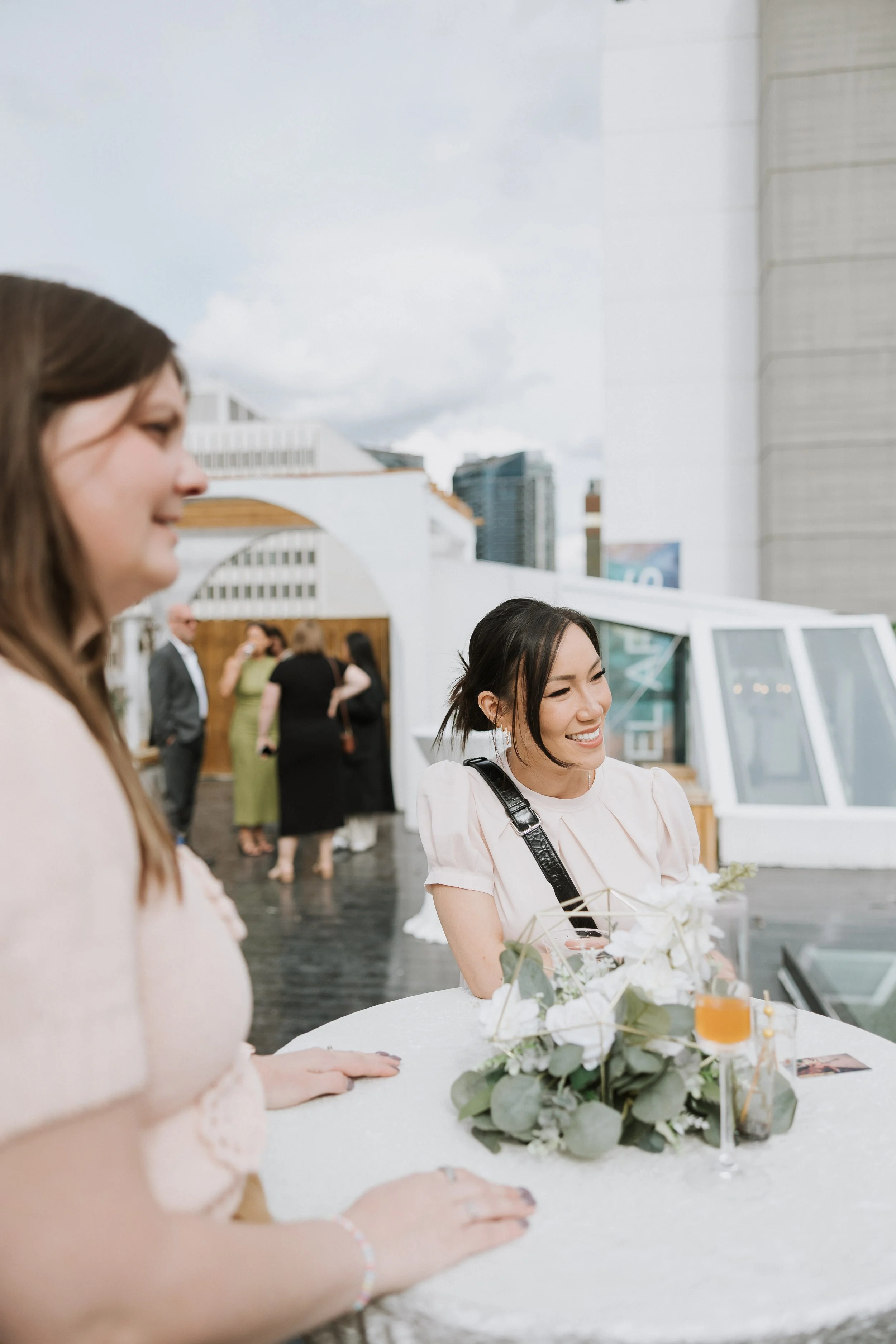 Three women smiling at a gathering, with a floral centerpiece on a table in front of them, and people in the background at an outdoor event.