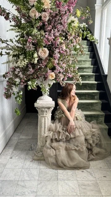 A woman in a beige ruffled dress sitting on the floor by a white pedestal with a large floral arrangement of pink and white flowers next to a staircase.