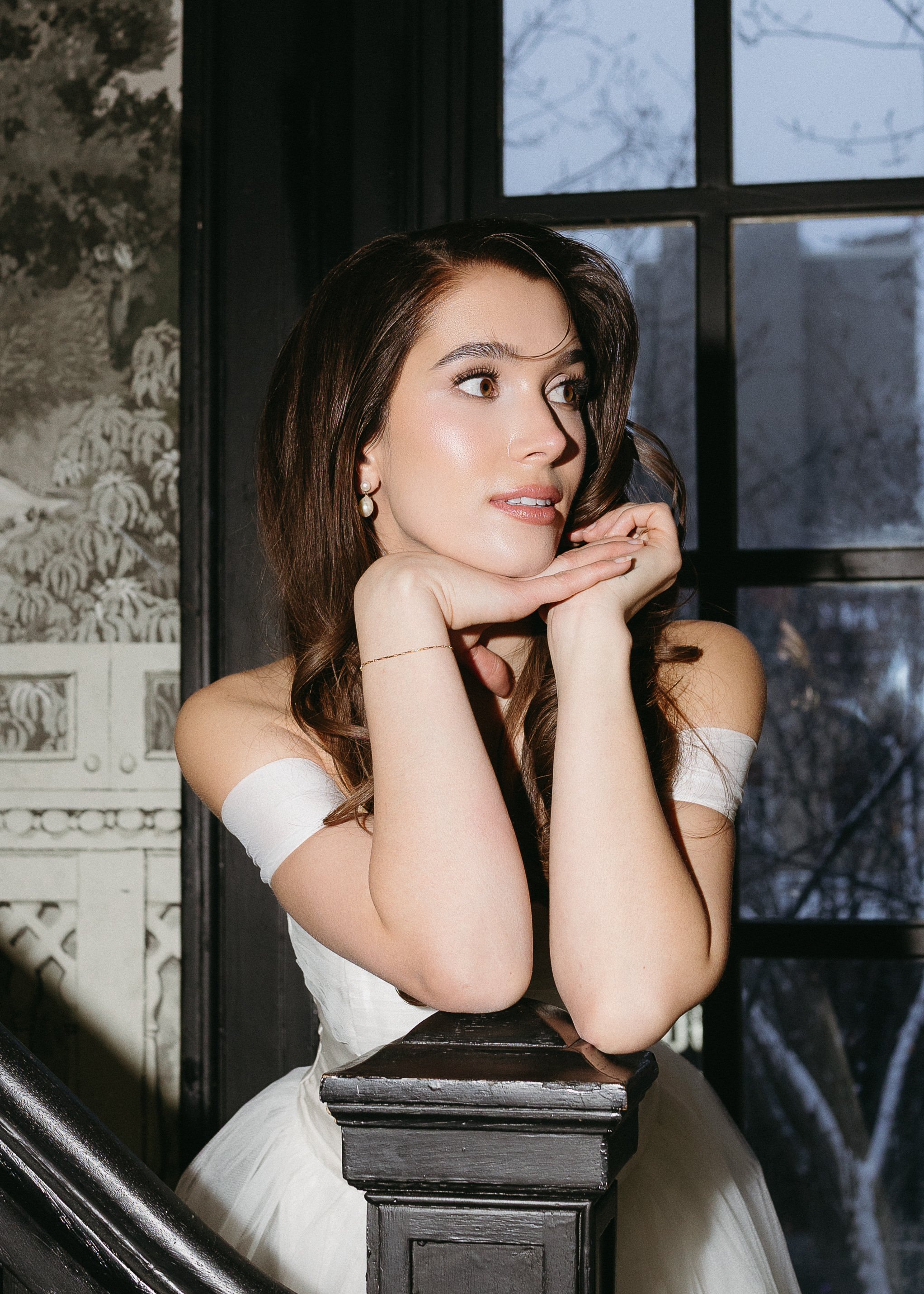 A woman with long brown hair, wearing pearl earrings and a white off-shoulder dress, poses resting her chin on her hands by a black wooden bannister with a window showing an outdoor scene with bare trees in the background.