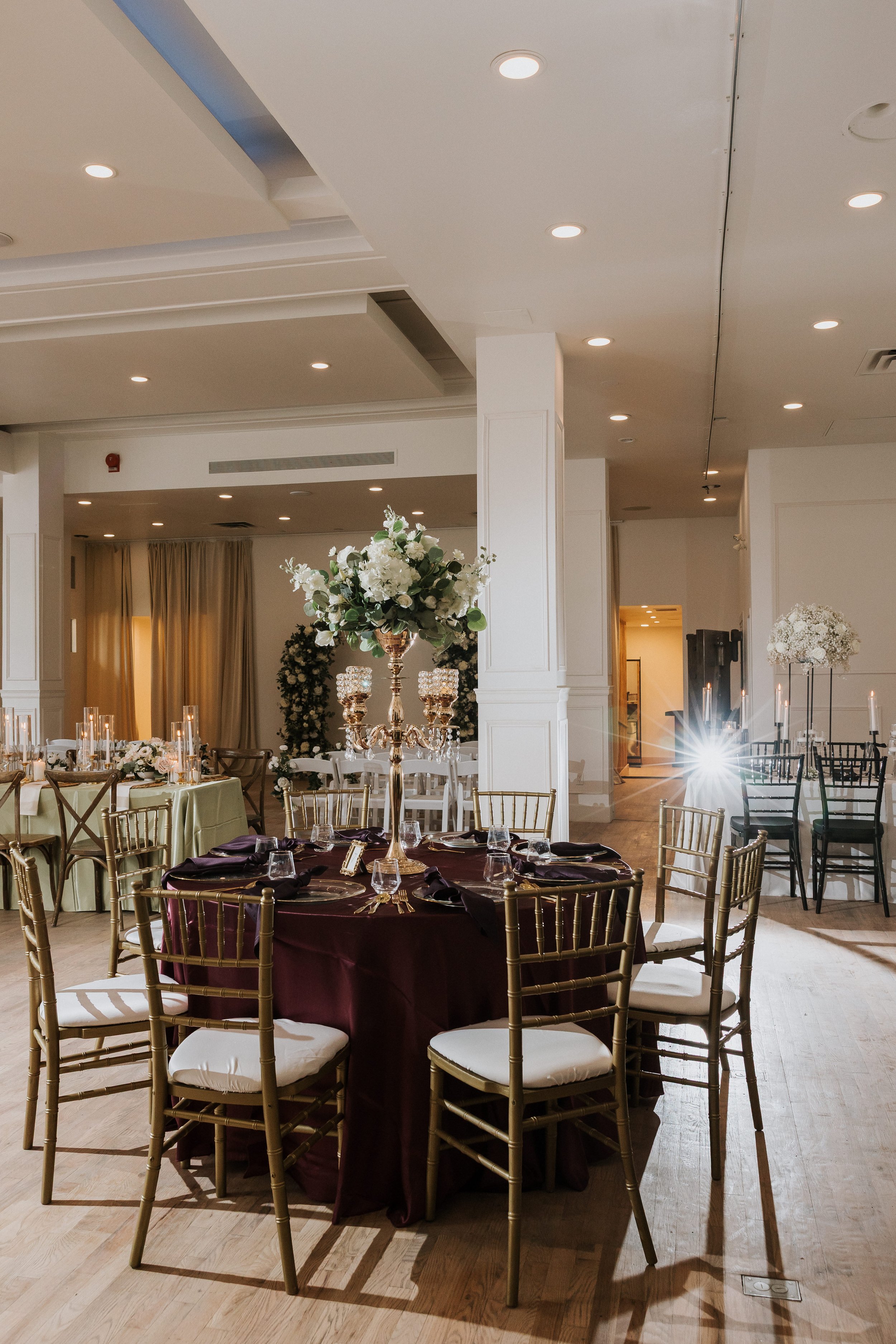Elegant event space decorated with floral centerpieces on tables, candleholders, and chairs set for a formal gathering or celebration in a bright room with high ceilings and warm lighting.