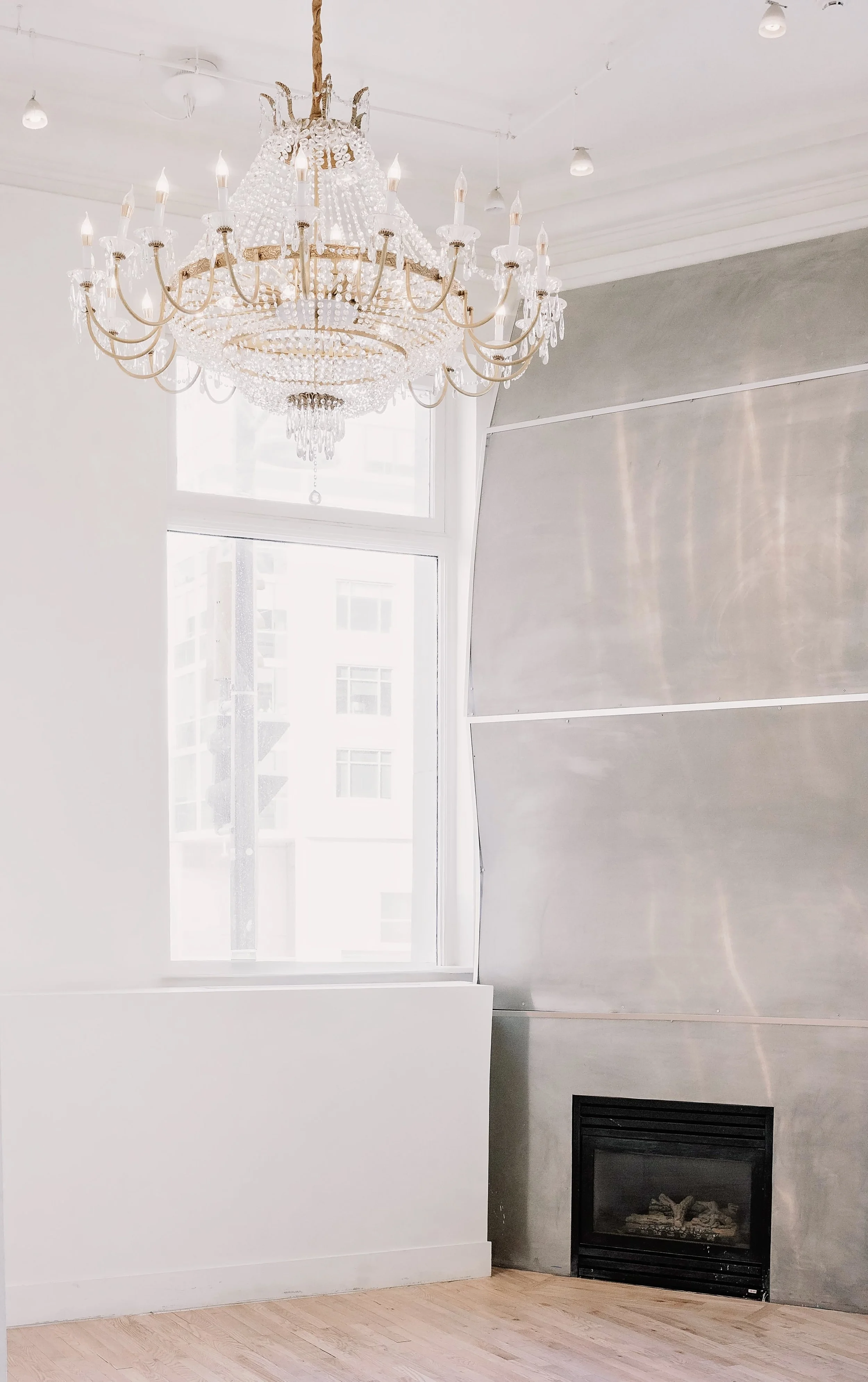 A modern living room corner with a large window, a chrome fireplace, and a grand crystal chandelier hanging from the ceiling.