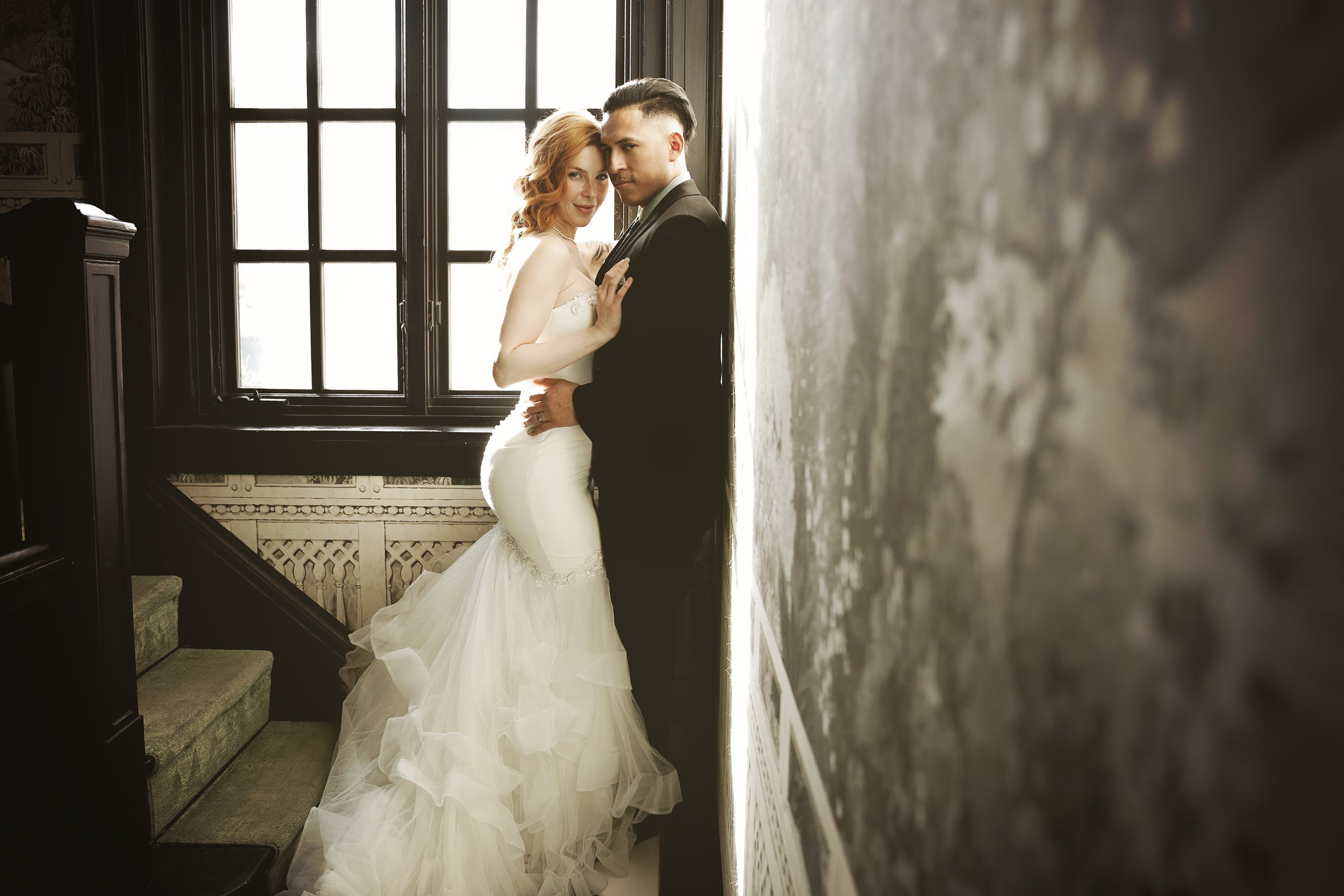 A bride and groom standing close together, leaning against a wall near a window with black trim. The bride has red hair and is wearing a white wedding dress, while the groom has dark hair and is wearing a black suit. They are indoors in a room with vintage-style decor.