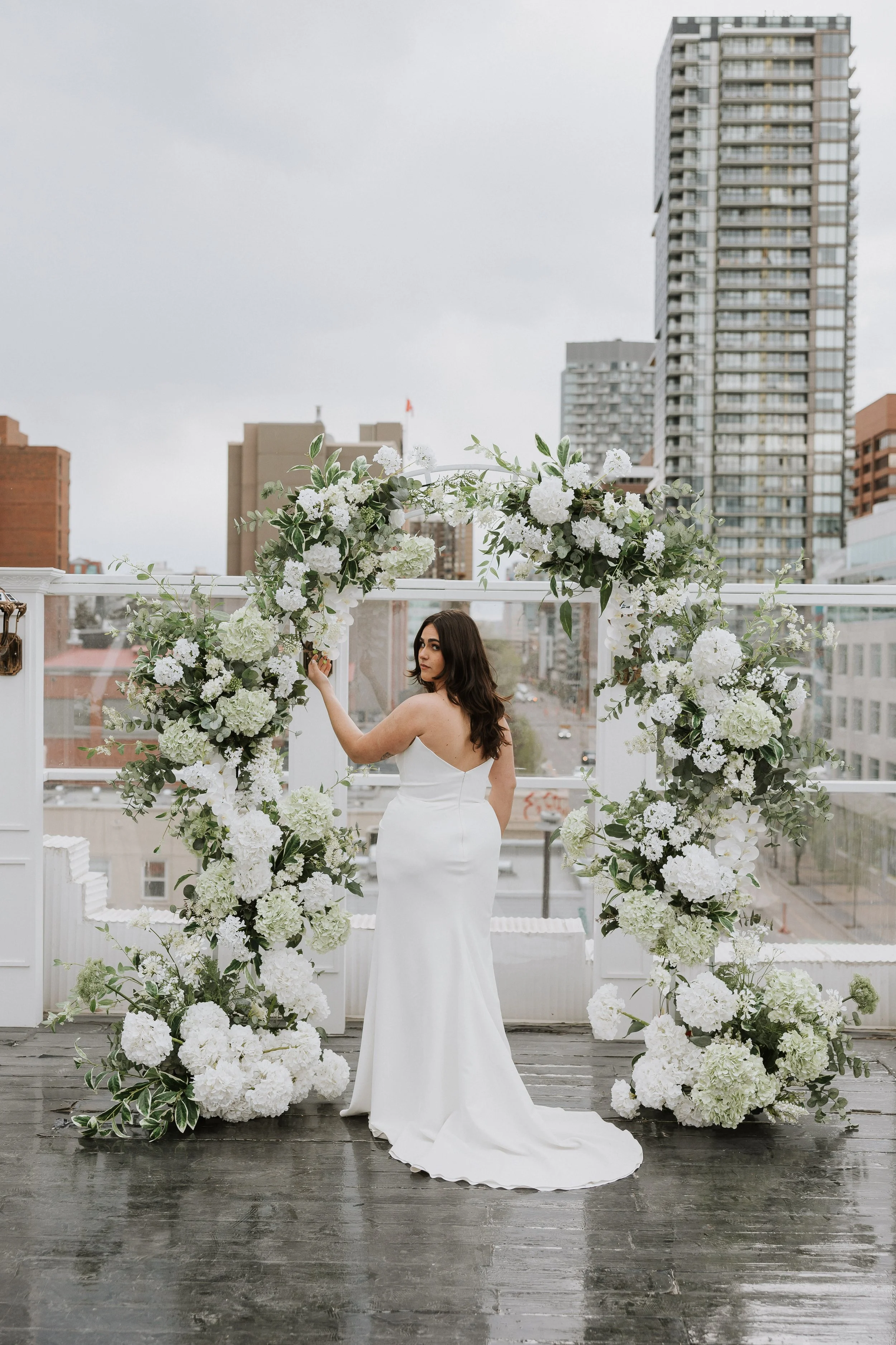 A bride in a white wedding dress with a train, standing under a white floral arbor on a rooftop with city buildings in the background.