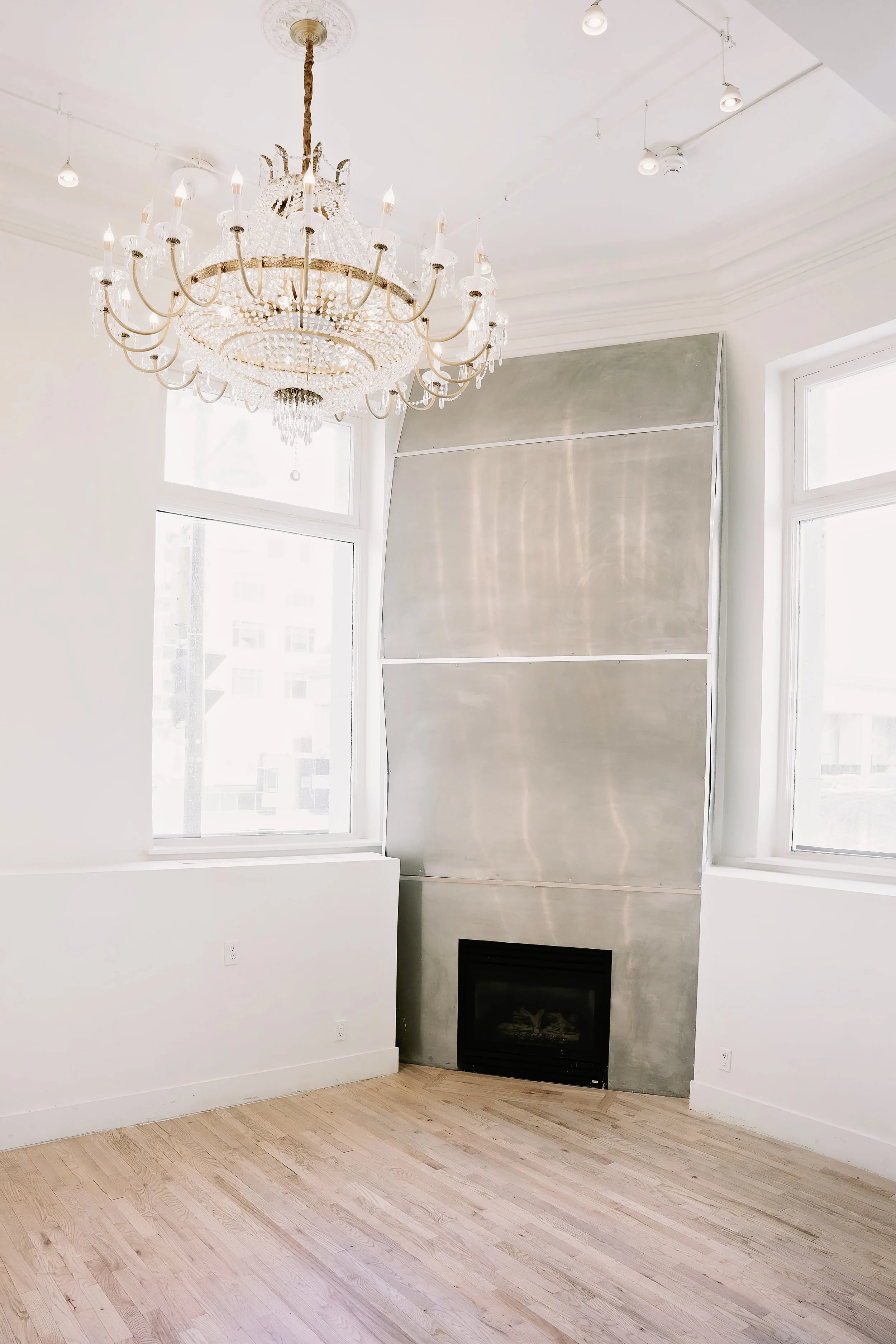 Empty living room with large windows, a modern fireplace on a gray wall, and a decorative chandelier hanging from the ceiling.