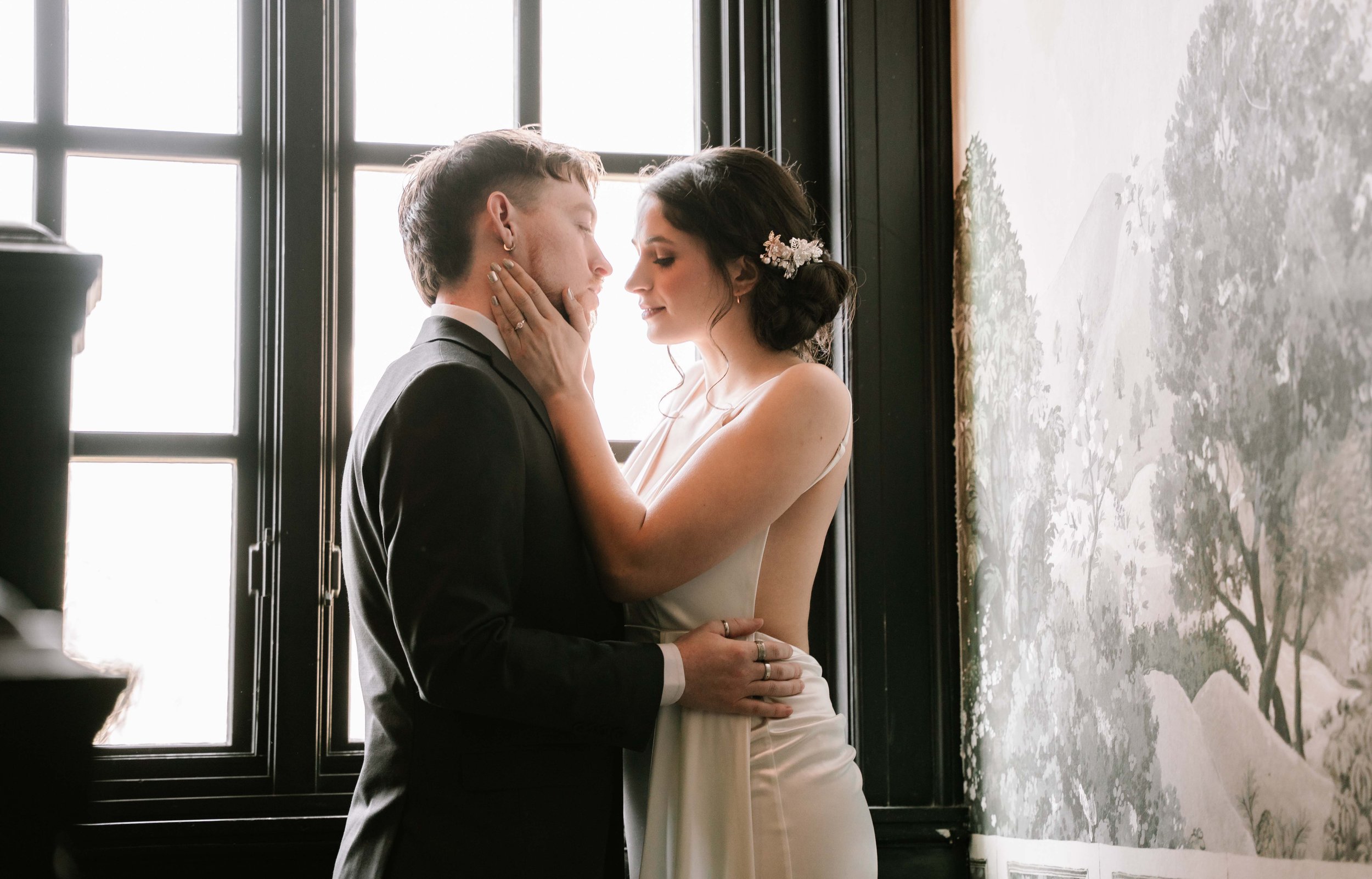 A bride and groom standing close to each other indoors, with the bride touching the groom's face, both with their eyes closed, in front of a large window and a textured wall with a mural of a tree.