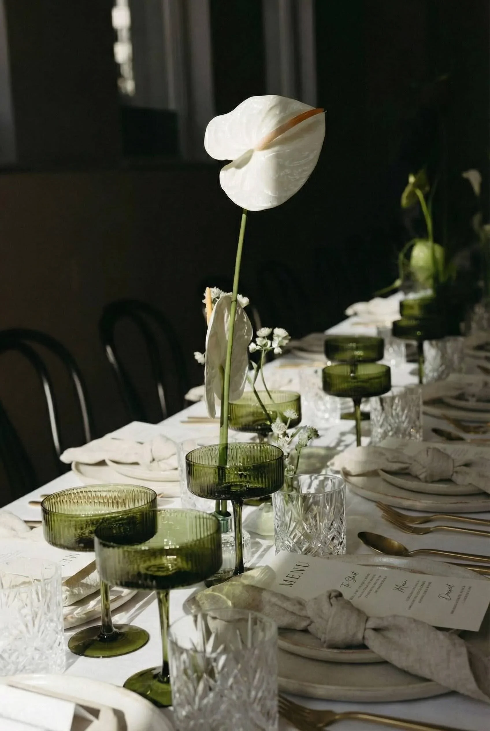 A long dining table decorated with white tablecloth, glassware, silverware, napkins, and green glass candle holders with white flowers and a single large white flower as centerpiece.