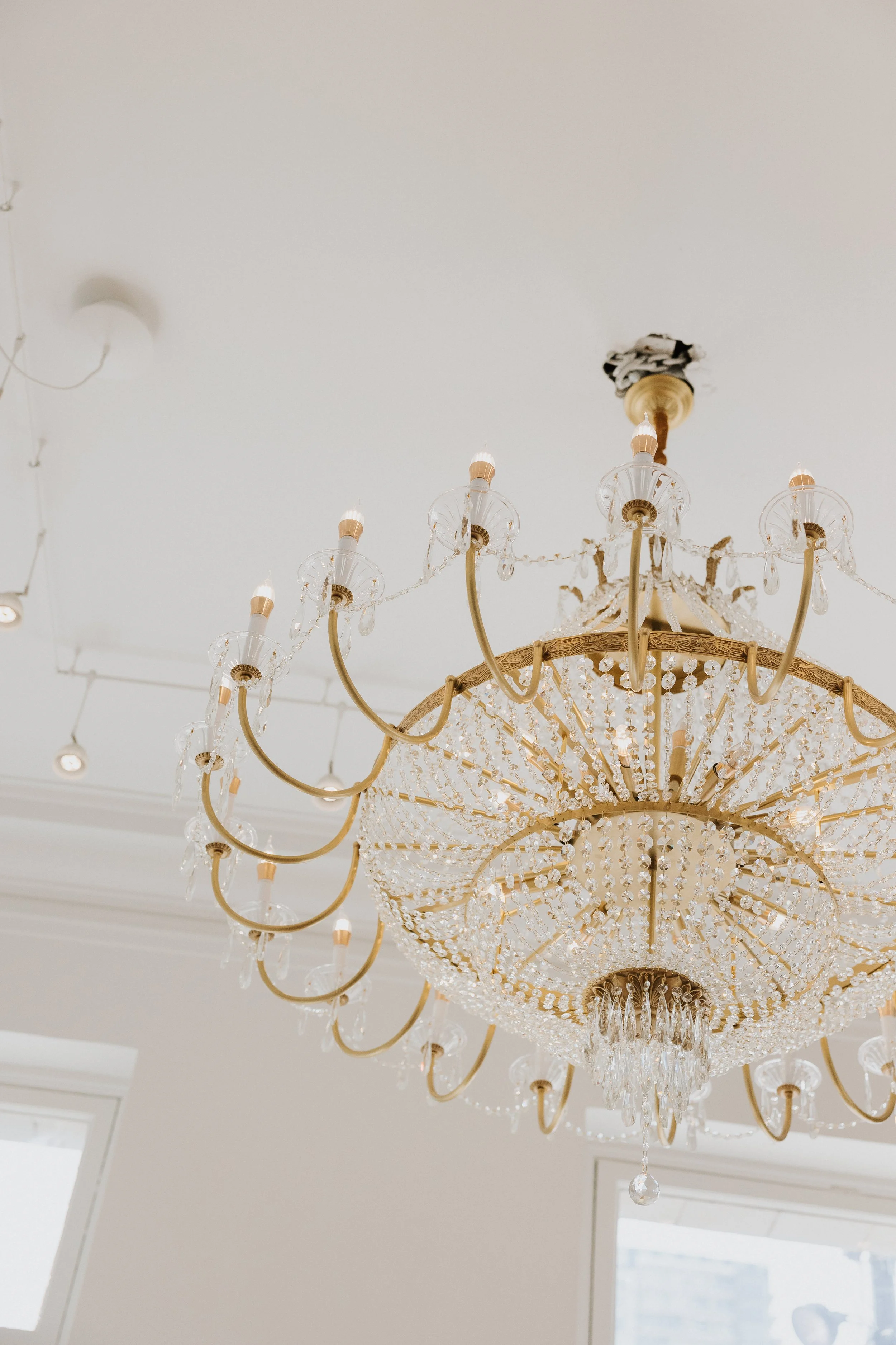 A large ornate chandelier with multiple tiers, crystal decorations, and candle-shaped light bulbs hanging from the ceiling of a bright room.