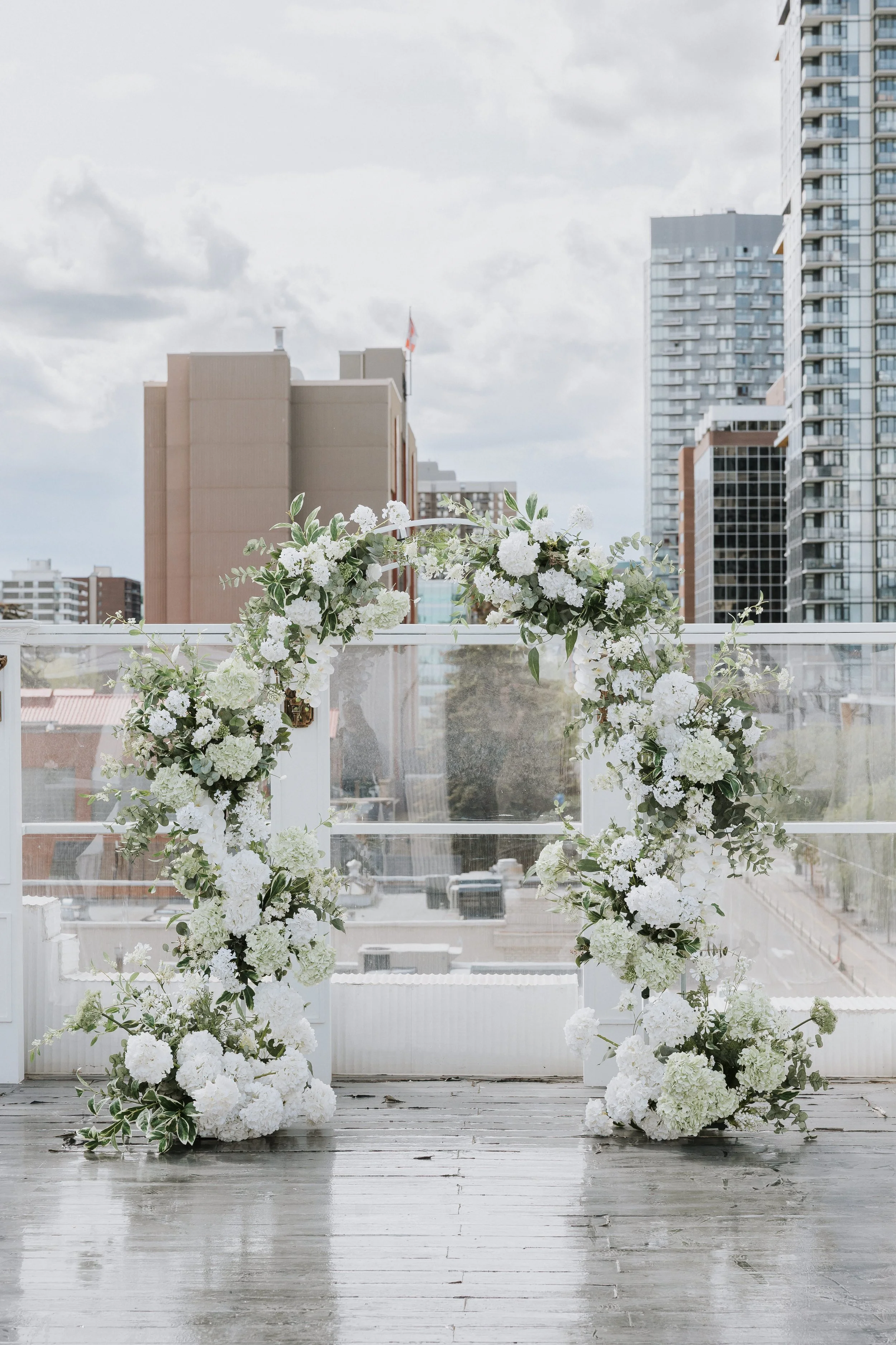 White floral arch decorated with white flowers and green foliage on a balcony, overlooking city buildings and a cloudy sky.