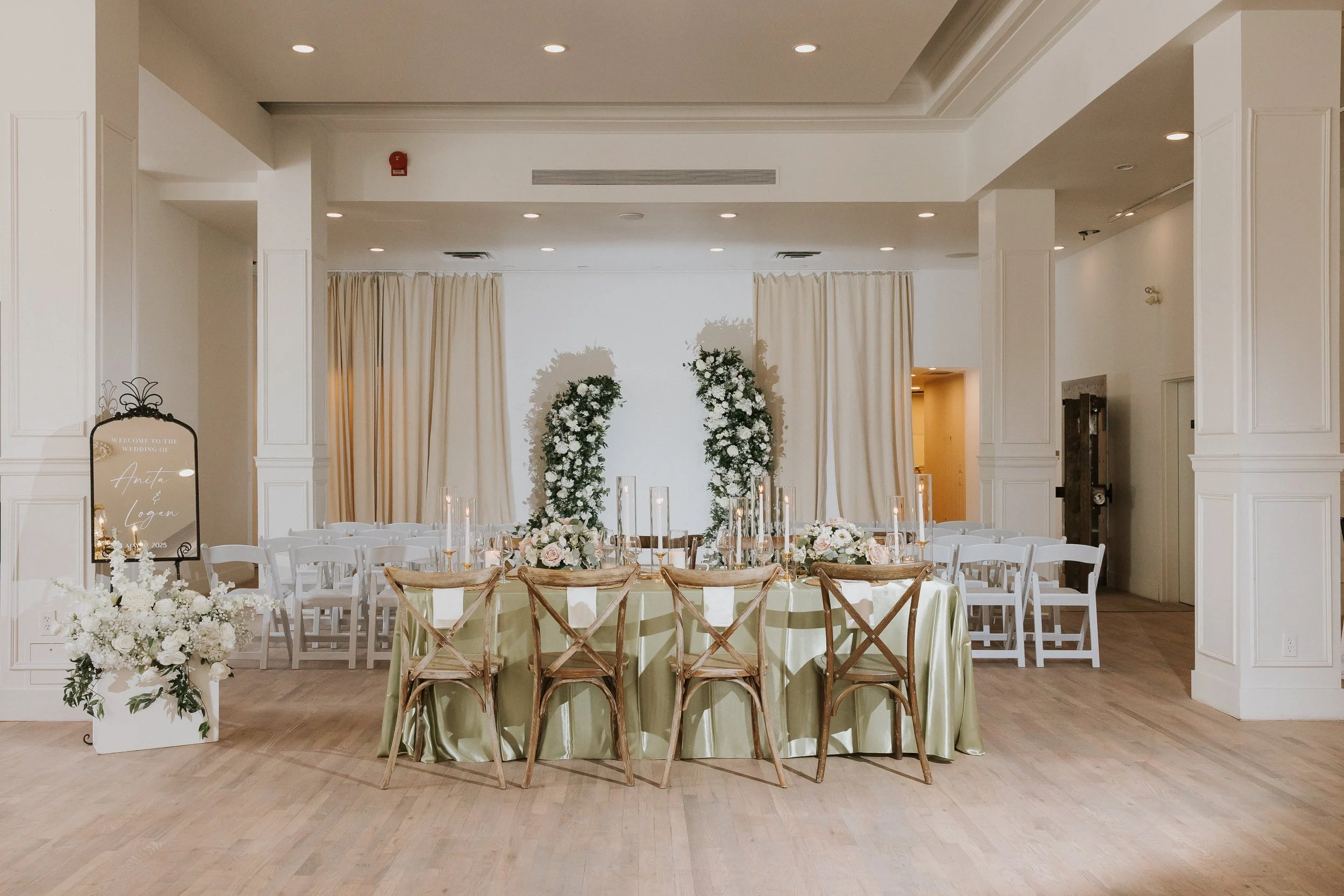 Elegant wedding reception setup with round table, floral centerpiece, candles, and white chairs in a bright, white room with curtains.