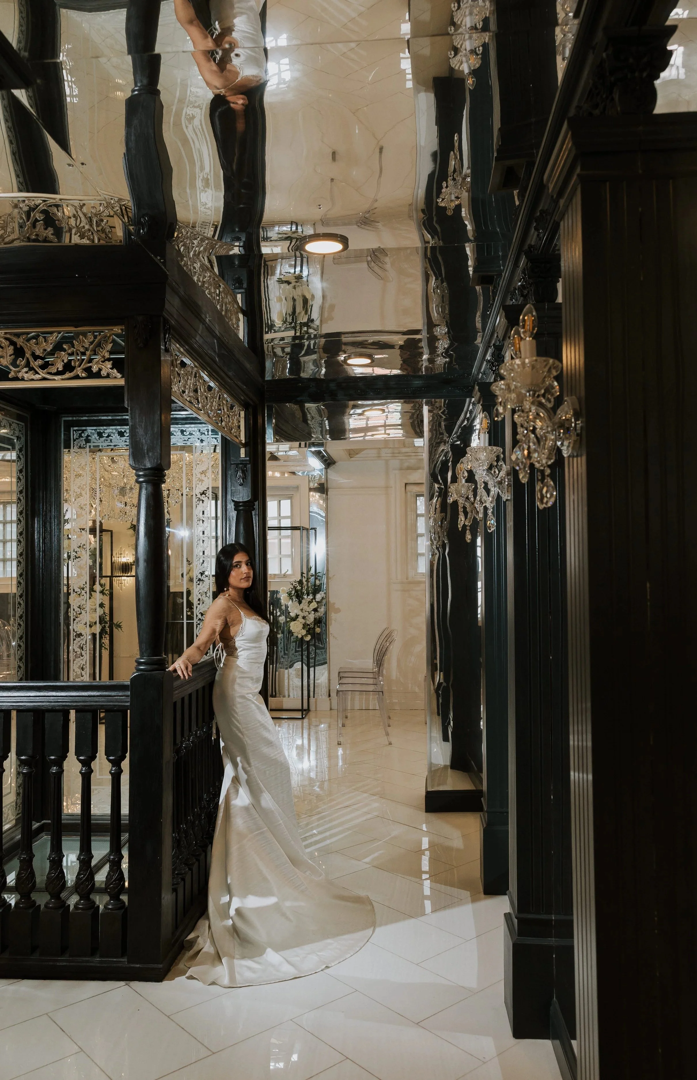 A woman in a white satin wedding gown standing in a luxurious, black and gold-adorned interior with chandeliers, mirrors, and elegant decor.