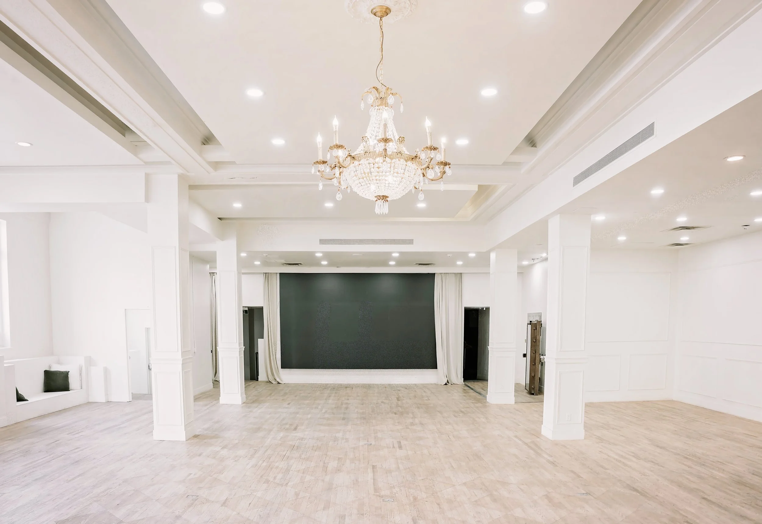 Elegant white hall with decorative molding, a crystal chandelier hanging from the ceiling, and a stage at the back with white curtains and a large black screen.