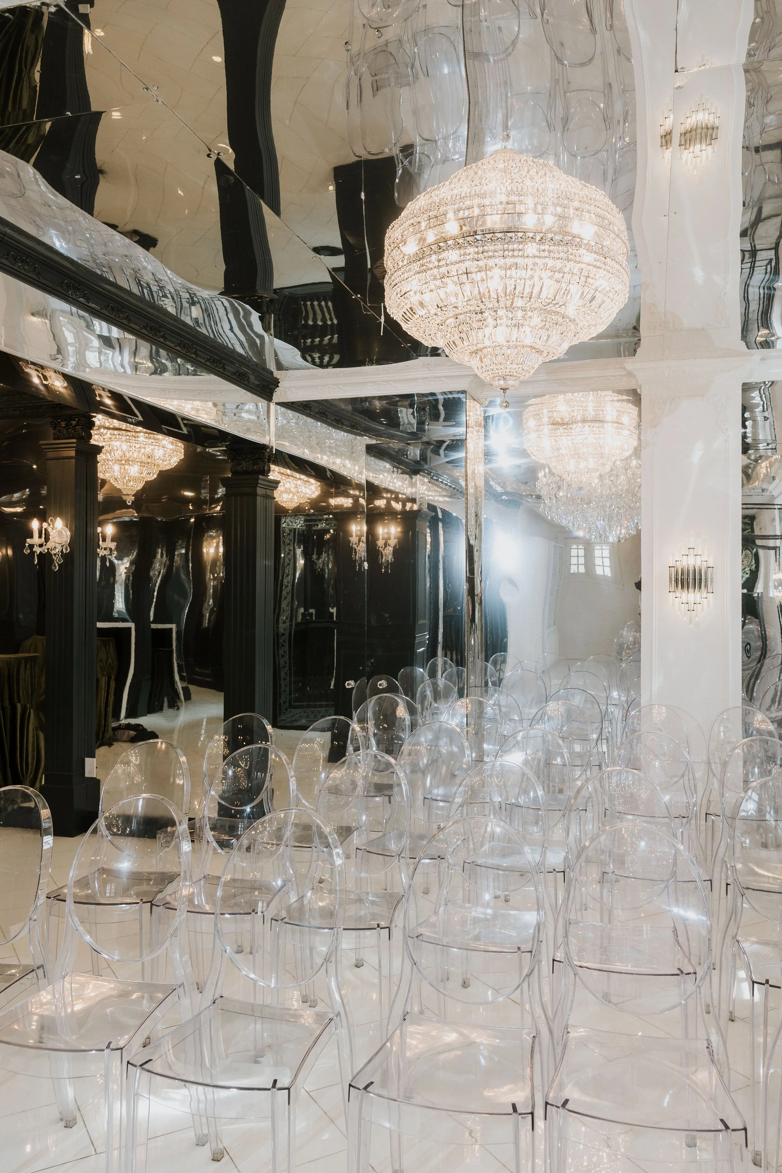 A luxurious interior with crystal chandeliers and transparent chairs arranged in rows, black columns, and reflective mirrored walls.