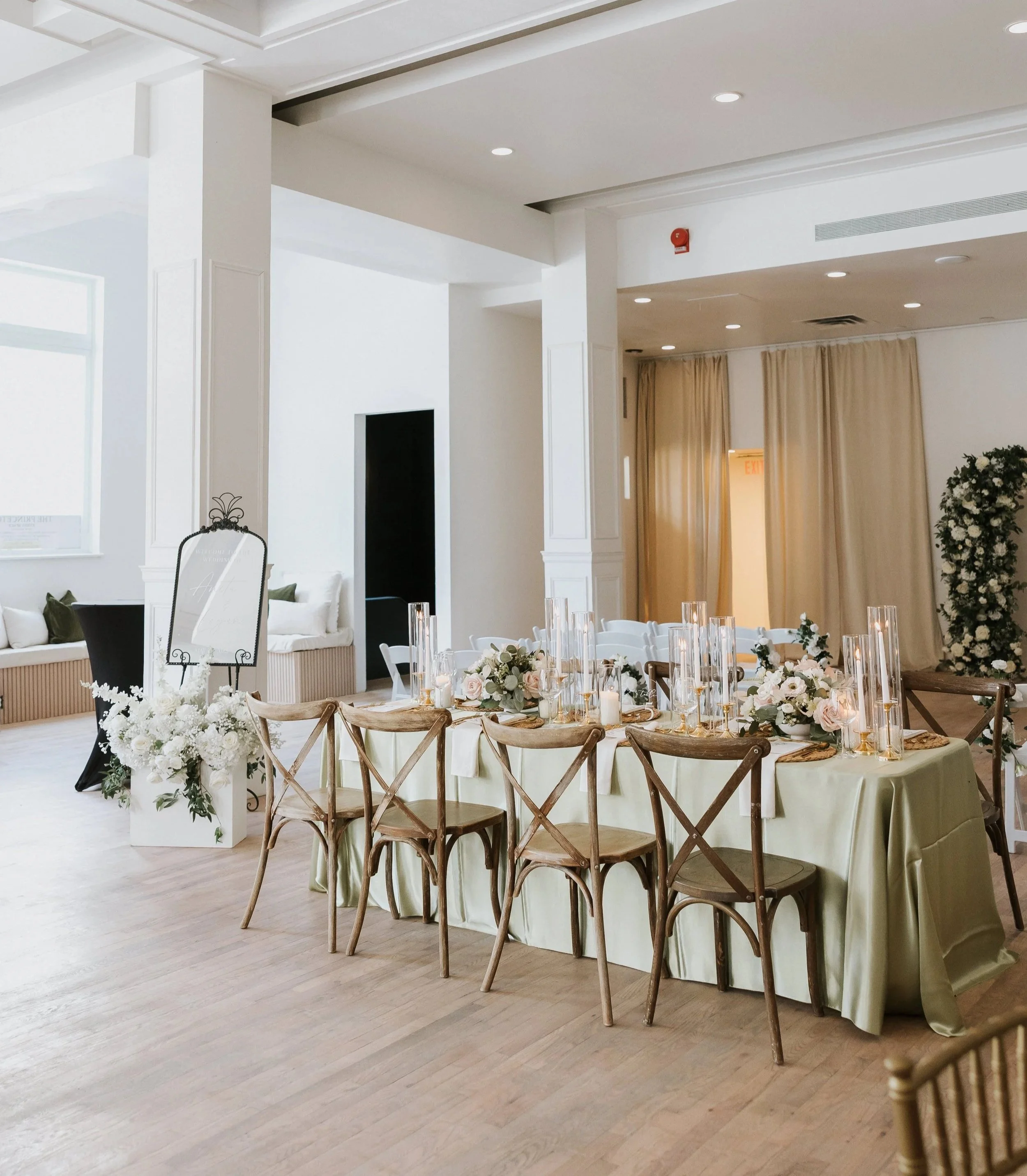 Elegant event space with round tables dressed in cream tablecloths, floral centerpieces, candles, and wooden cross-back chairs. The background features white walls, beige curtains, and warm lighting, creating a sophisticated atmosphere.