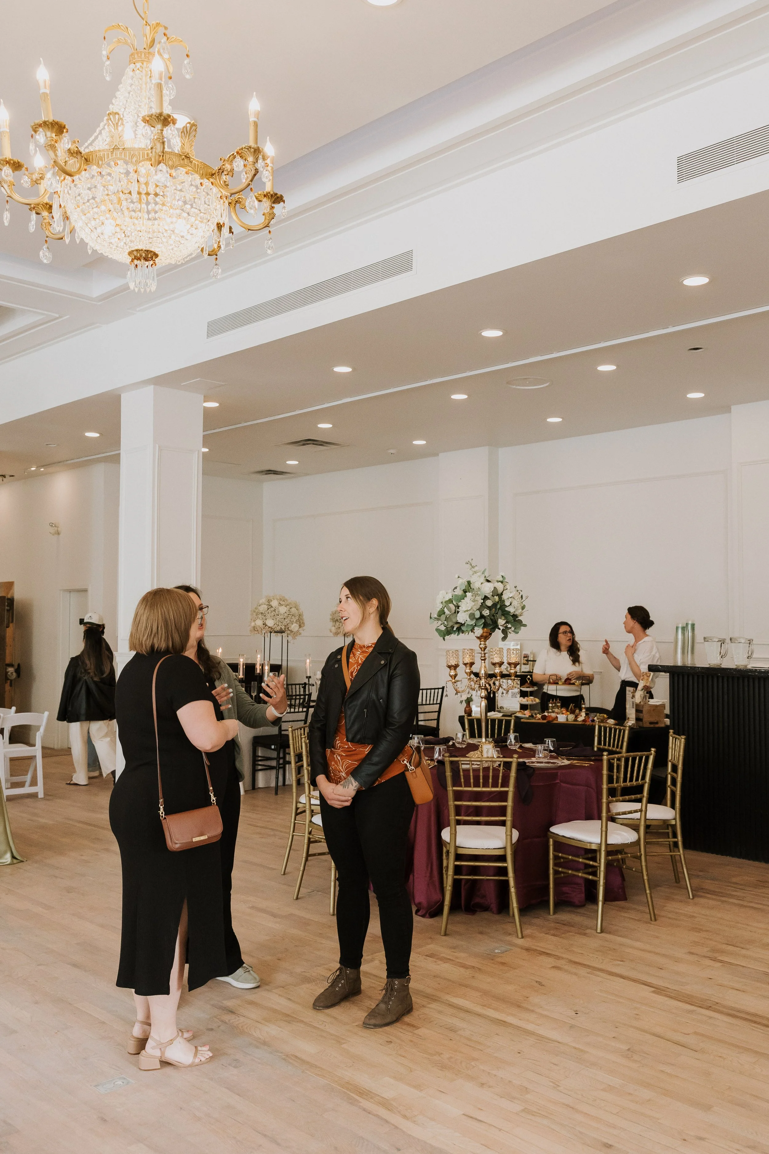 Group of women conversing at a formal event with decorated tables, floral arrangements, and a chandelier in an elegant banquet hall.