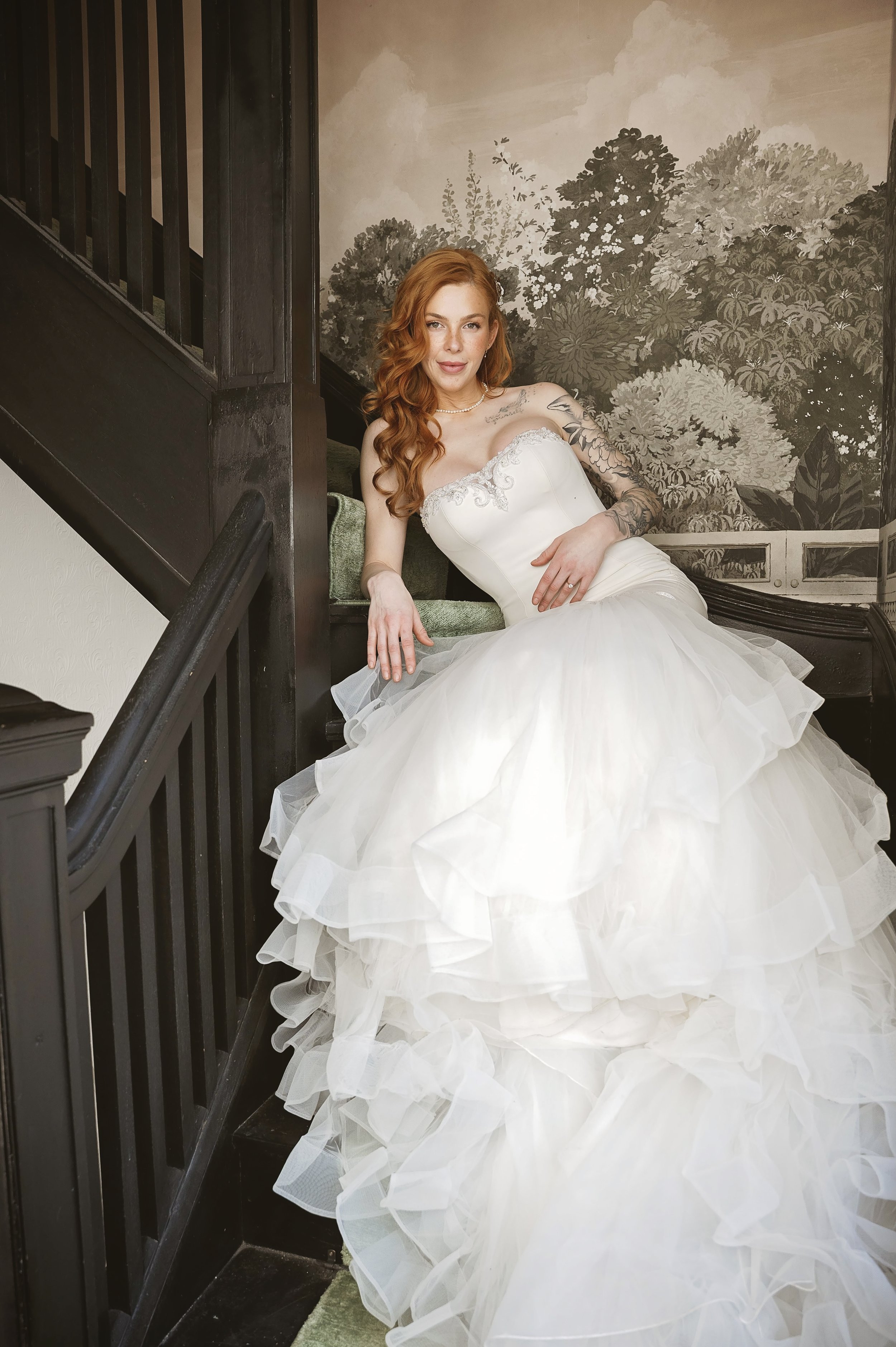 A woman with red hair in a white wedding dress sitting on a staircase with a floral mural in the background.