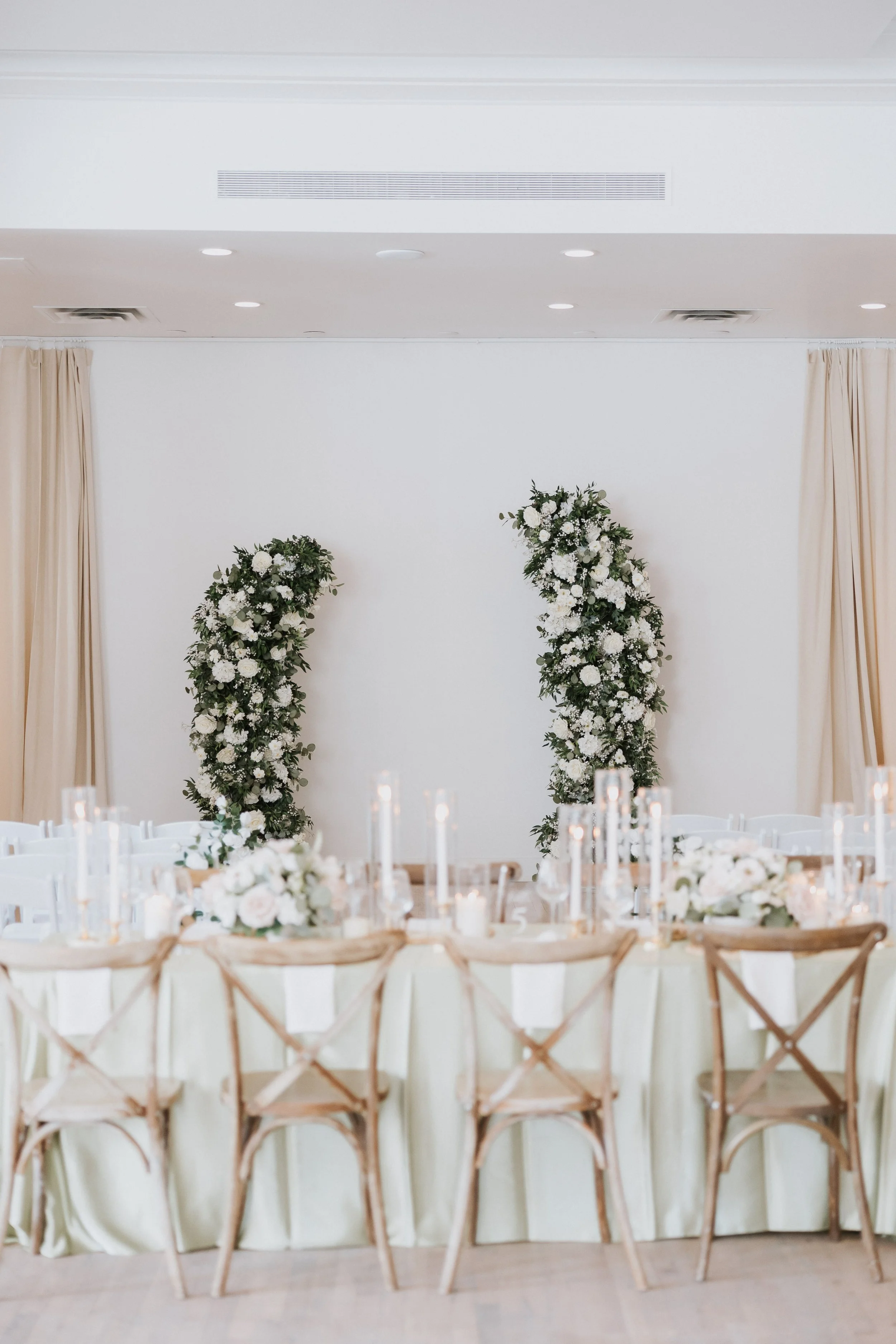 Wedding reception setup with round tables, white tablecloths, floral centerpieces, and wooden chairs, in front of a decorated wall with floral arrangements and candle decor.