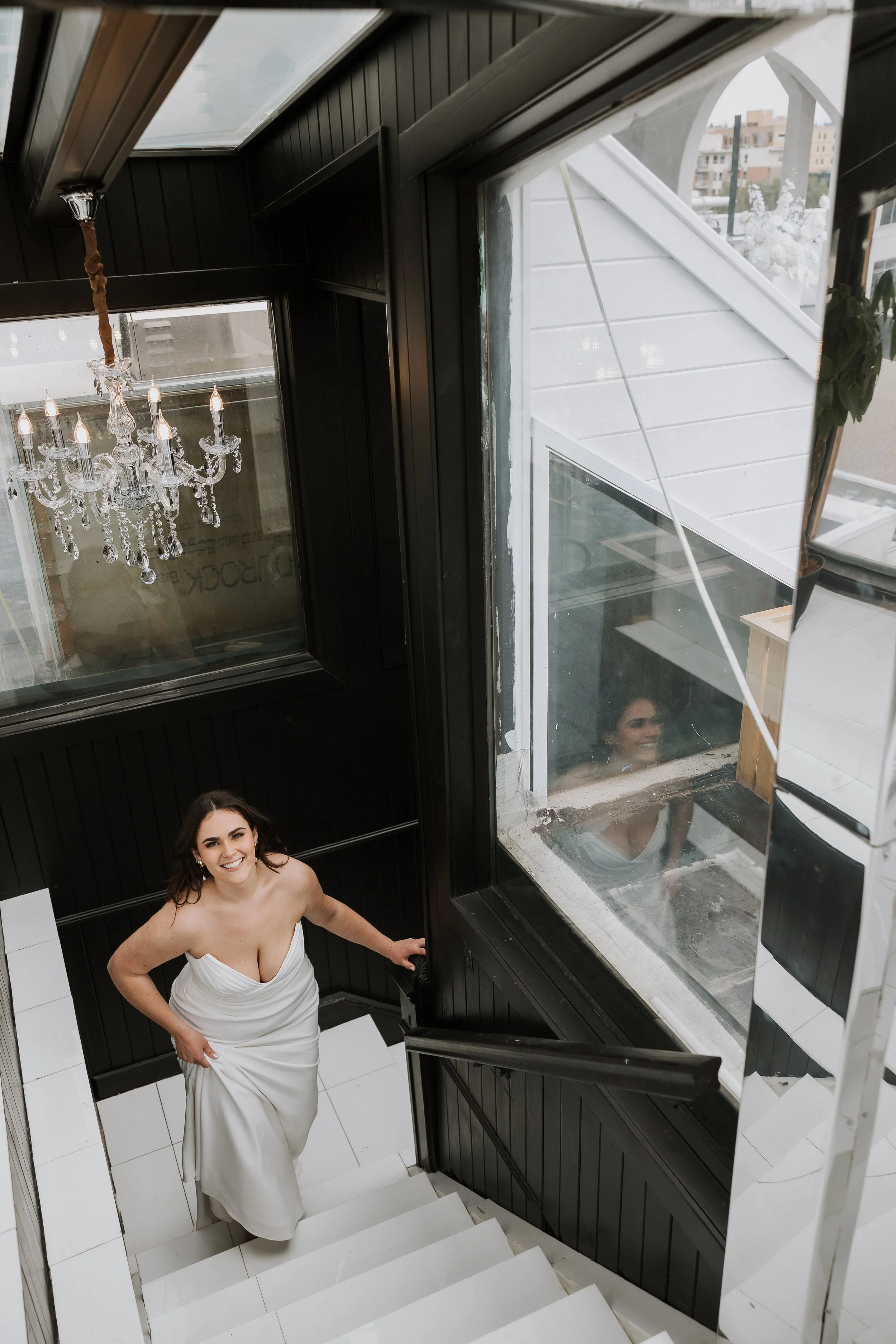 A woman wearing a strapless white wedding dress standing on a staircase, smiling and looking up at the camera. The staircase is black and white with a chandelier hanging above. There are windows and city buildings visible in the background.