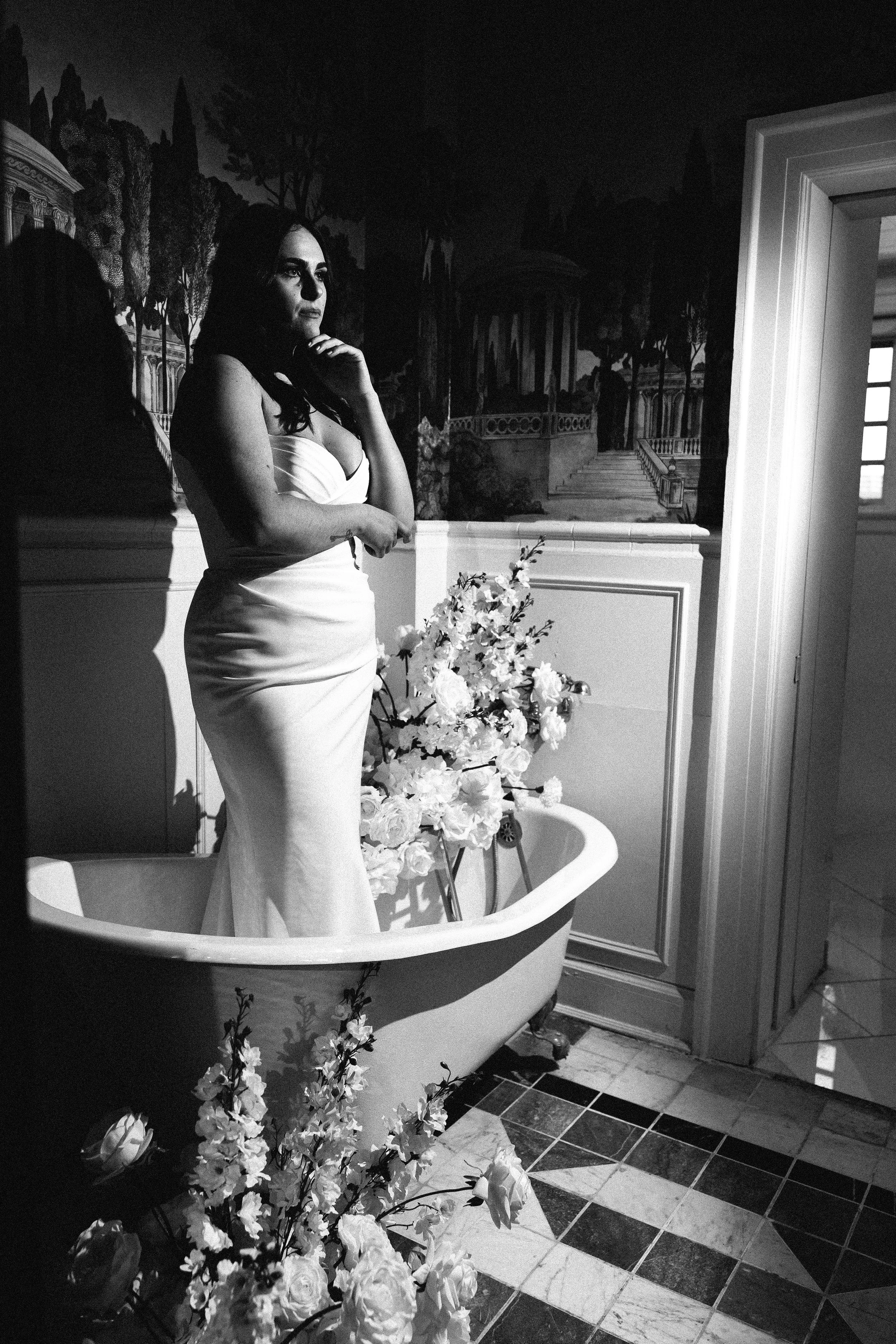 A woman in a strapless white gown standing beside a vintage bathtub with floral decorations inside and around it, with a background mural of a garden scene on the wall.