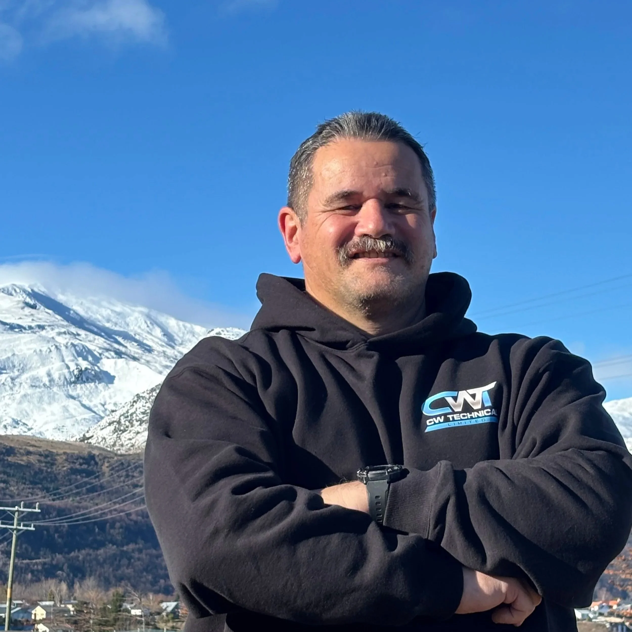 Close-up of a smiling man with salt-and-pepper hair and beard, wearing a dark CW Technical hoodie, standing outdoors with snow-capped mountains and blue sky in the background.