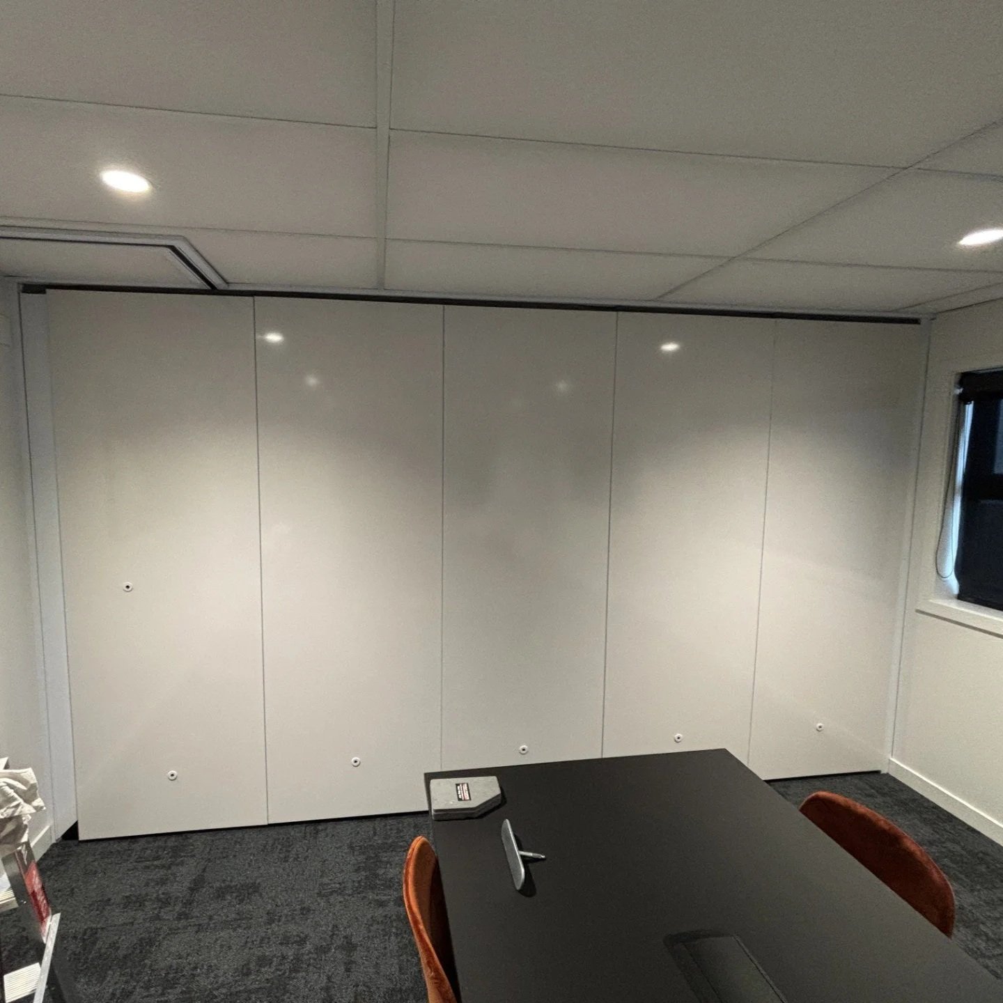 Office meeting room with a white wall, a black table, and four chairs, with a window on the right and ceiling lights.