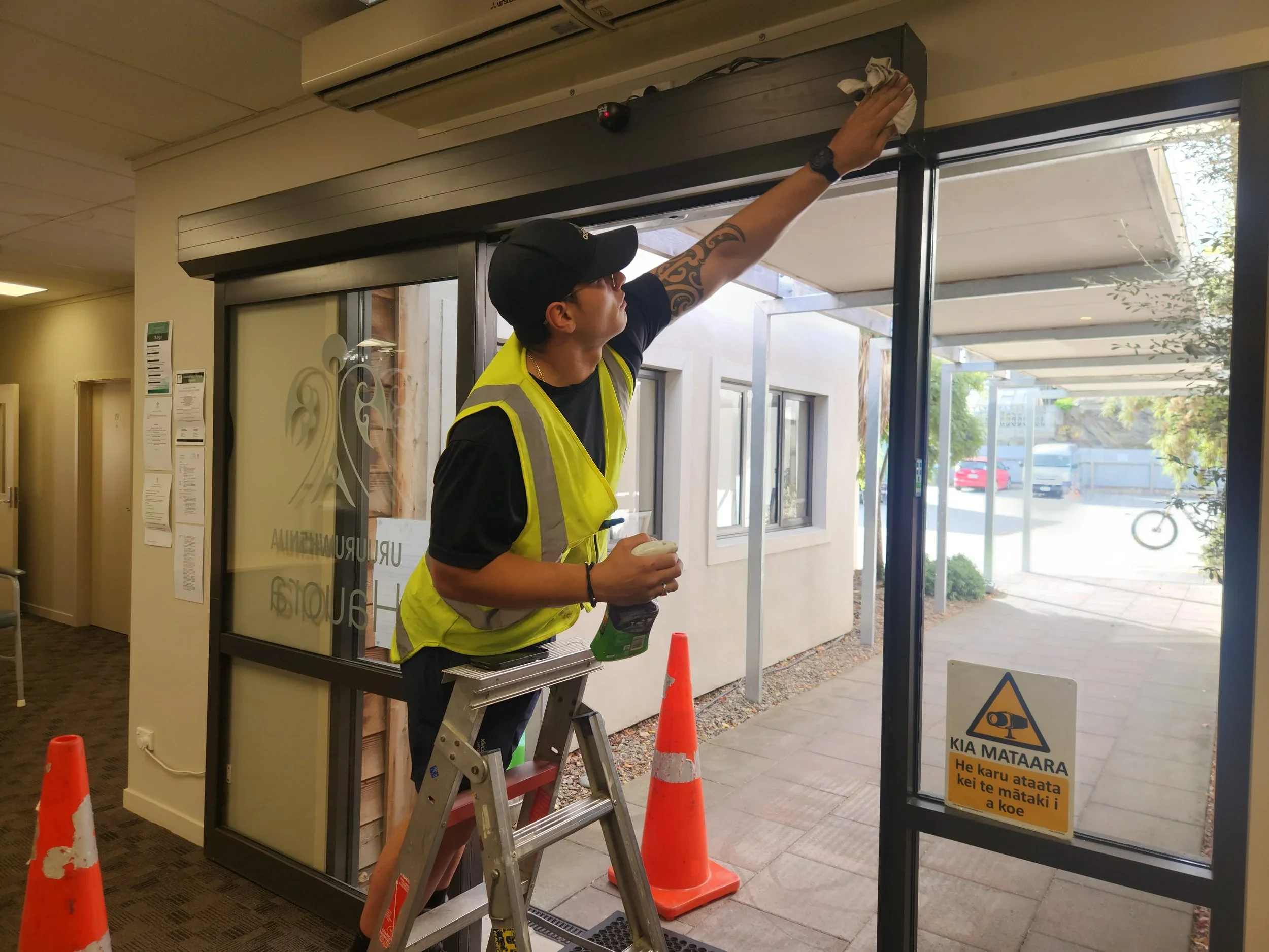 A person wearing a black cap and yellow reflective vest is cleaning a glass door with a cloth while standing on a small step ladder inside a building with orange cones on the floor outside the door. There are warning signs on the door, and the outside area shows parked vehicles and trees.