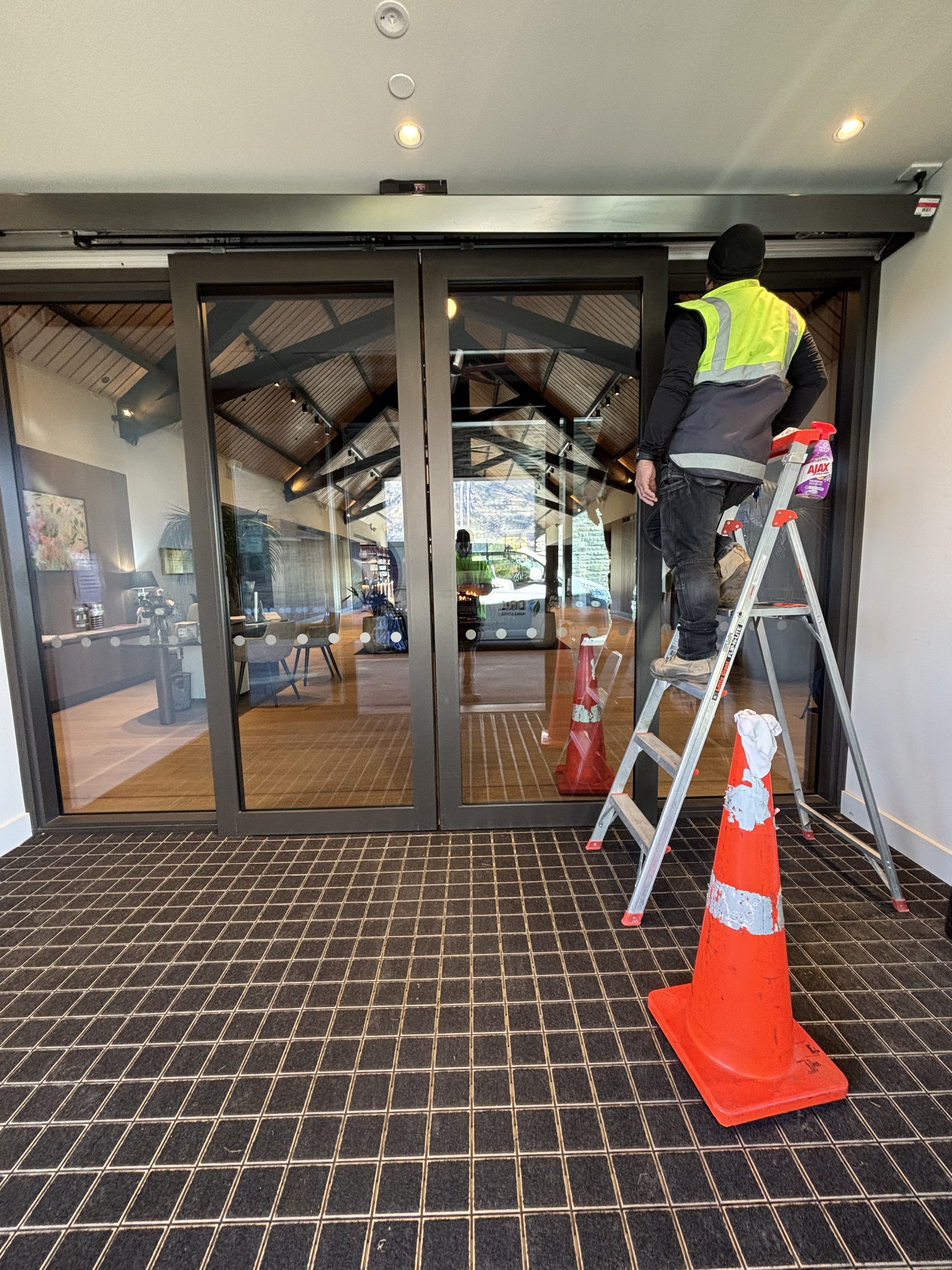 Worker in a black beanie and reflective vest standing on a ladder cleaning or inspecting a glass door at the entrance of a building, with orange traffic cones placed on a black tile floor outside.