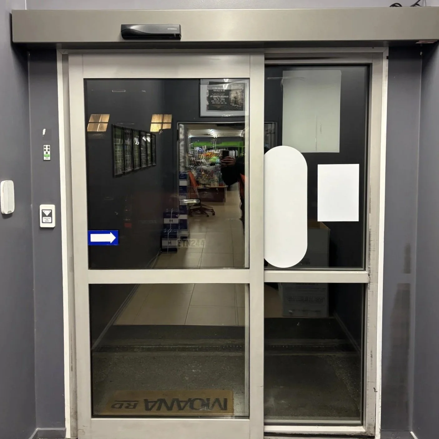 Automatic sliding glass door at a building entrance with a blue arrow sign indicating the direction to the right, a white rectangular sticker, and a gray wall surrounding the door.
