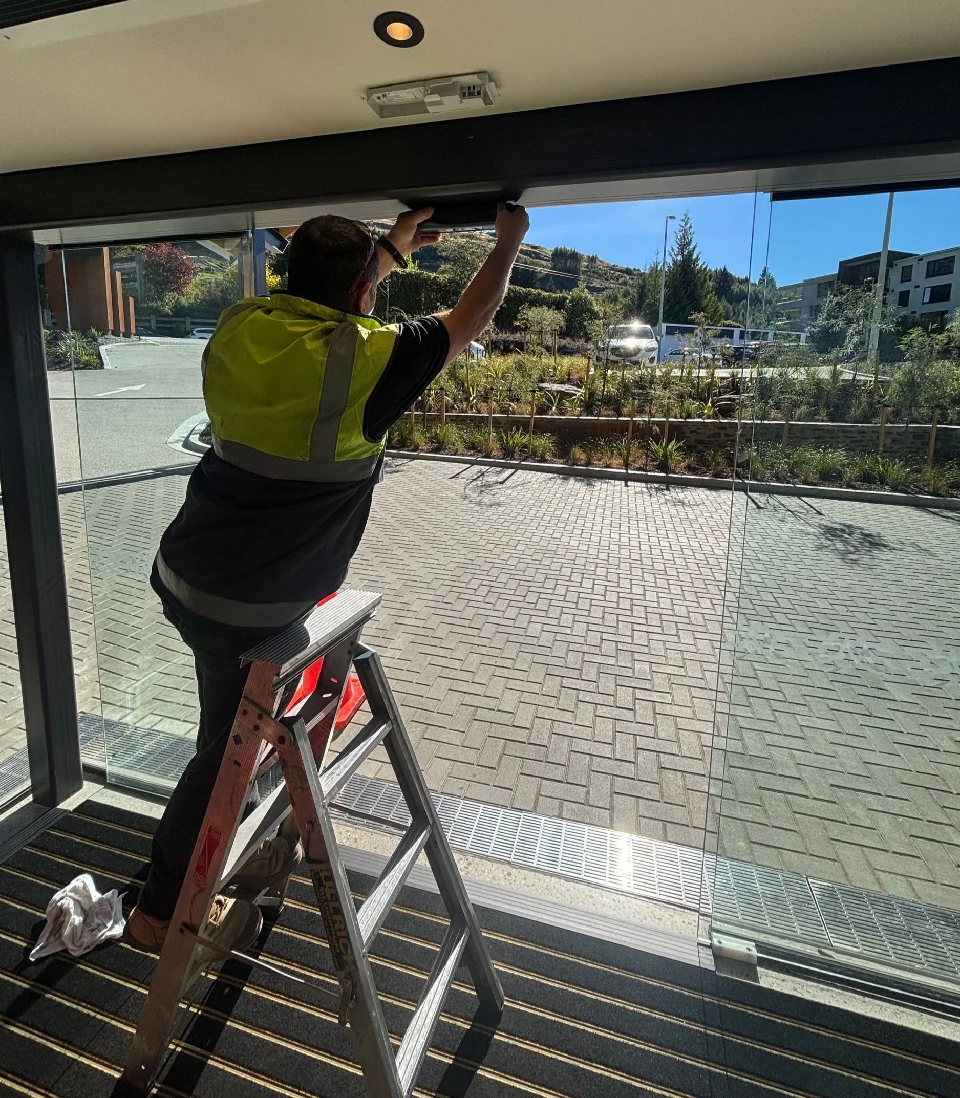 A person standing on a ladder inside a building, working on a ceiling or light fixture near a large glass window, with a view of a parking lot, plants, and residential buildings outside on a sunny day.