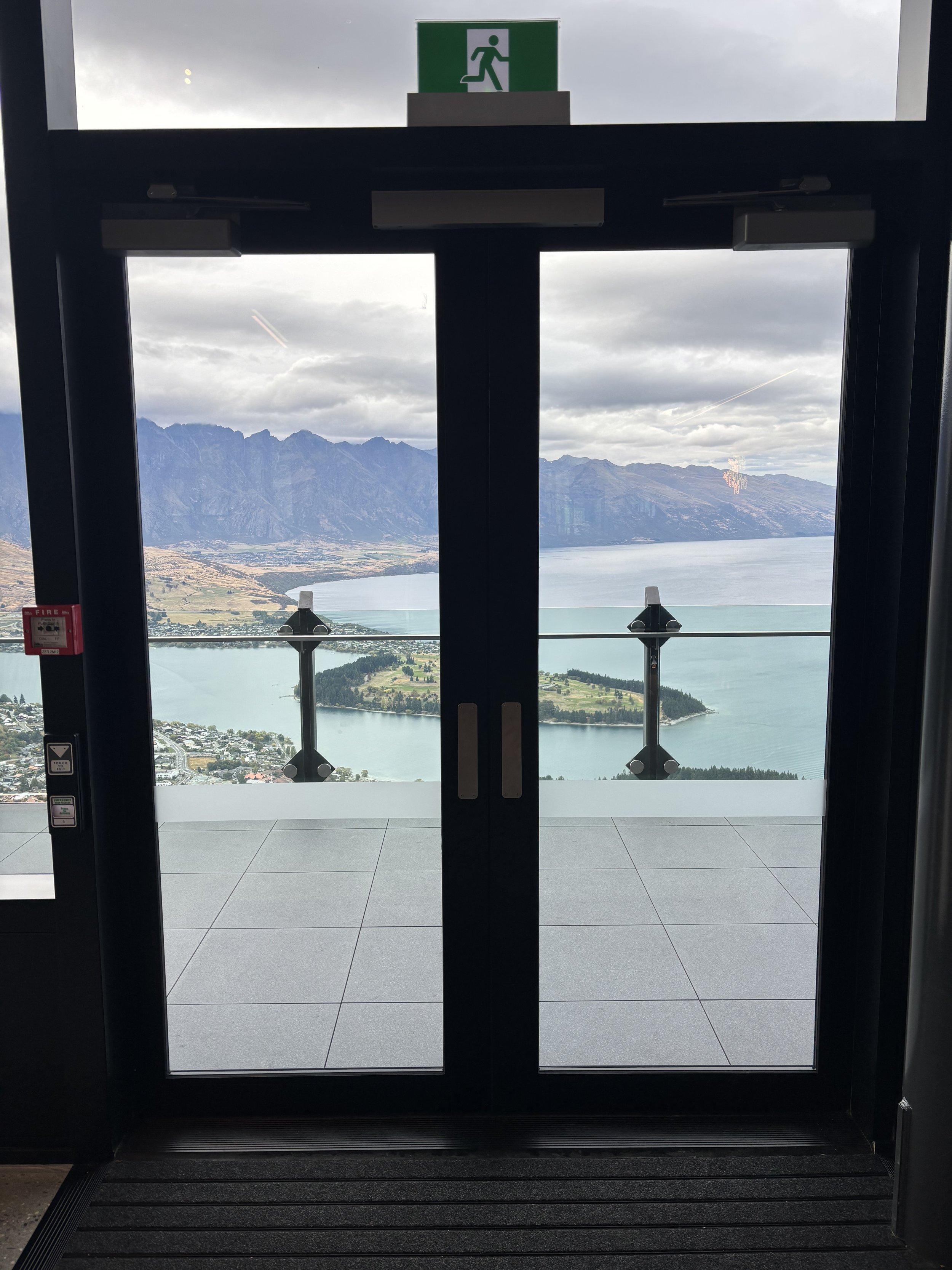 View through glass doors at an airport, showing a scenic landscape of mountains, lake, and town under cloudy sky.