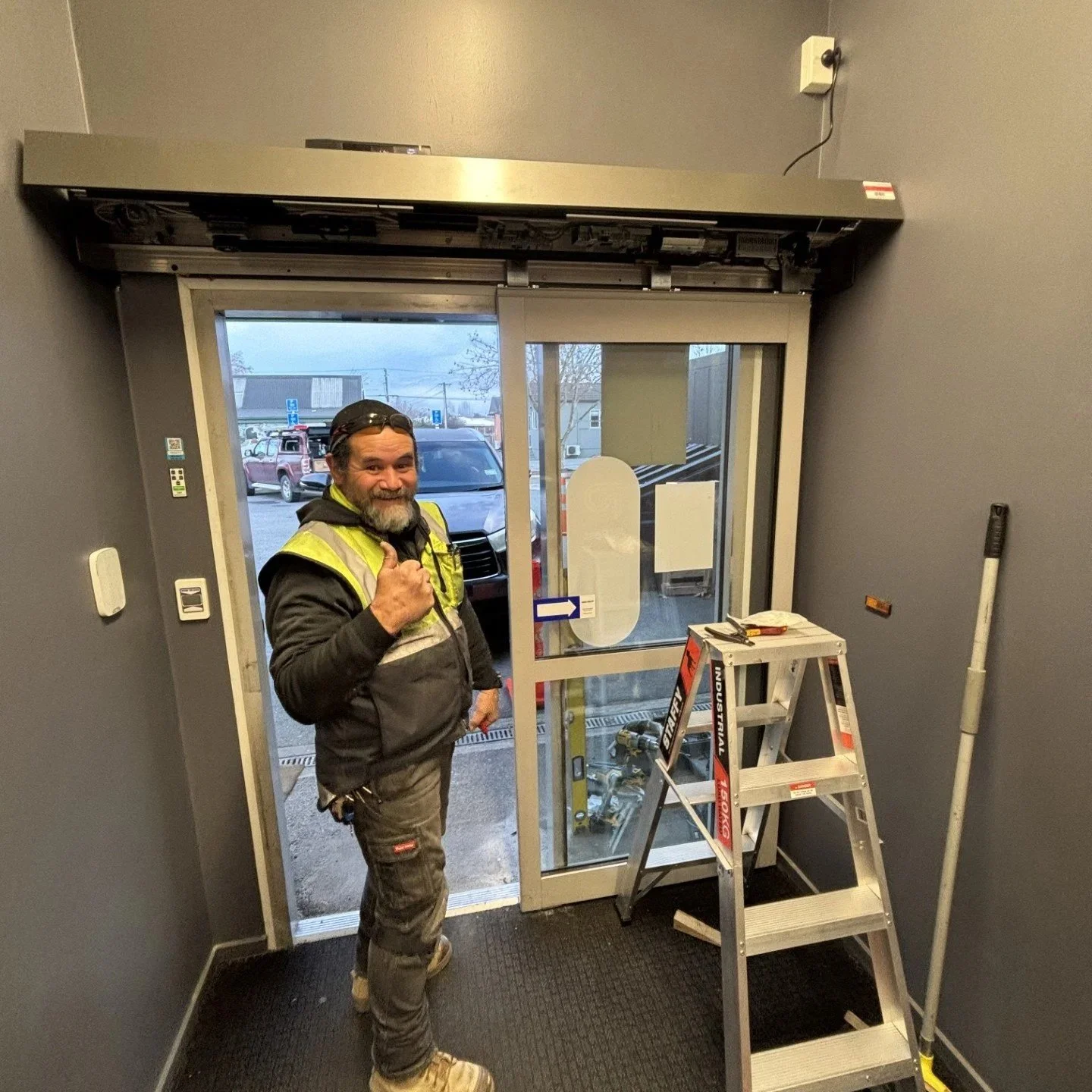A man in construction work attire, including a high-visibility vest, standing inside a building near an entrance door, giving a thumbs-up gesture, with a step ladder and construction tools nearby.